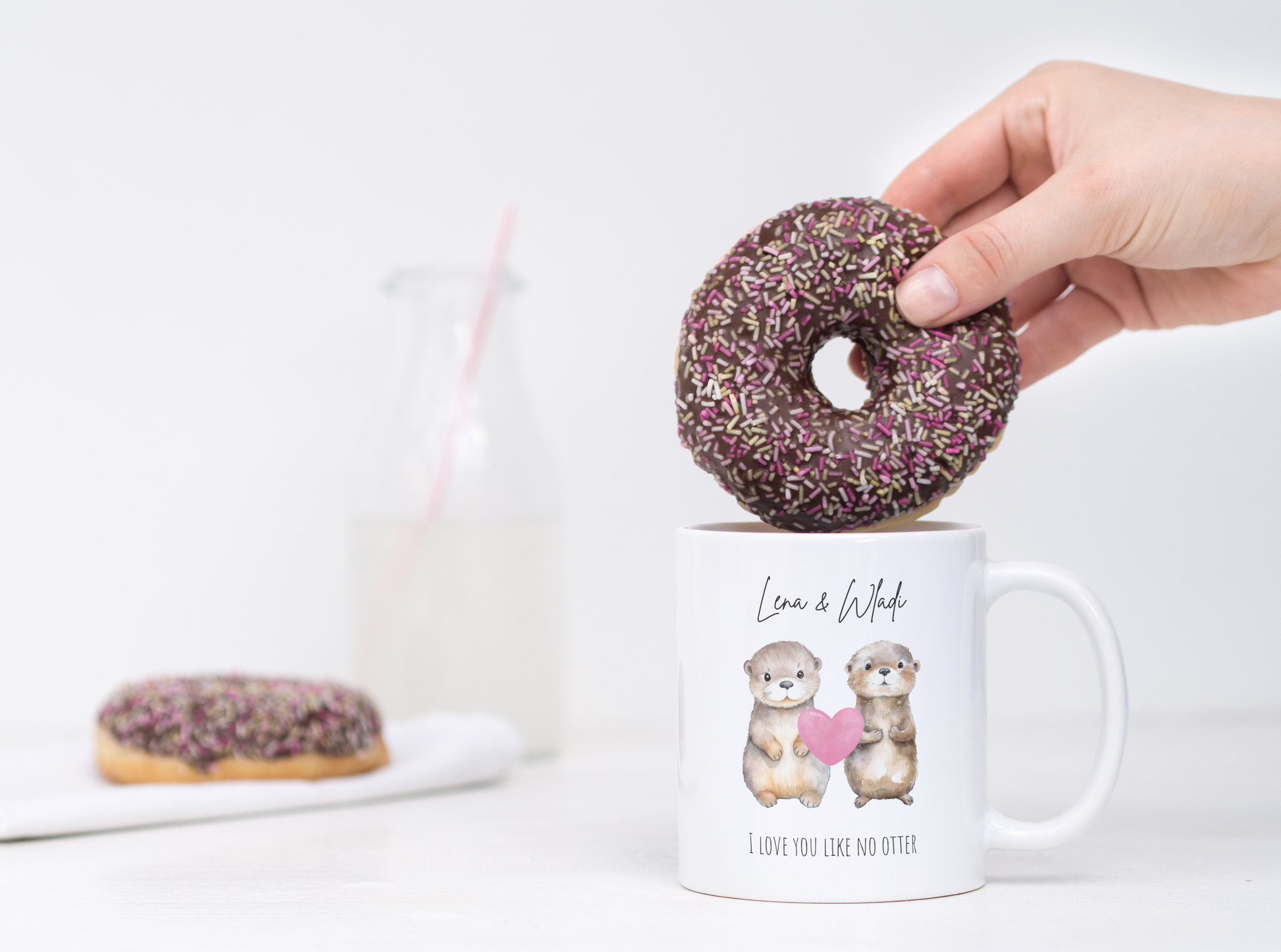 Ein Schokodonut wird in eine weiße Tasse mit zwei süßen Ottern und der Aufschrift "Lena & Wladi" und "I love you like no otter" getunkt