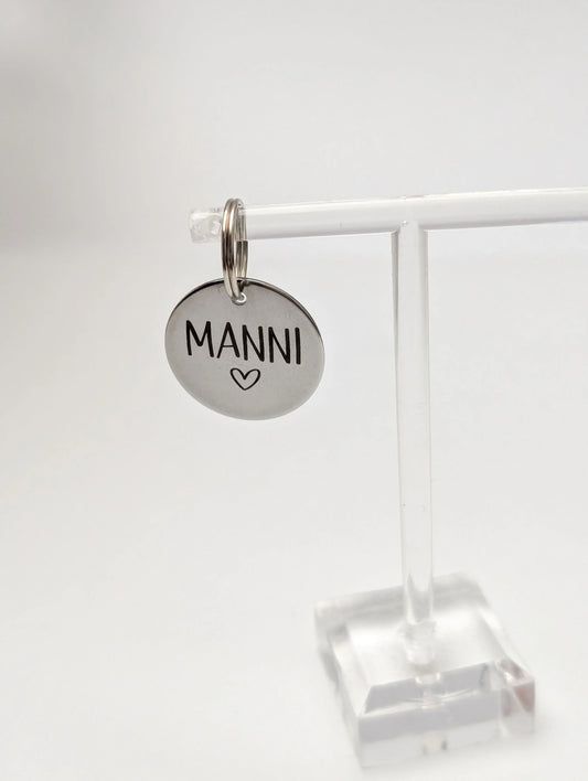 Silberne Hundemarke mit Gravur "Manni" und einem Herz hängt an einem durchsichtigen Schmuckständer vor weißem Hintergrund