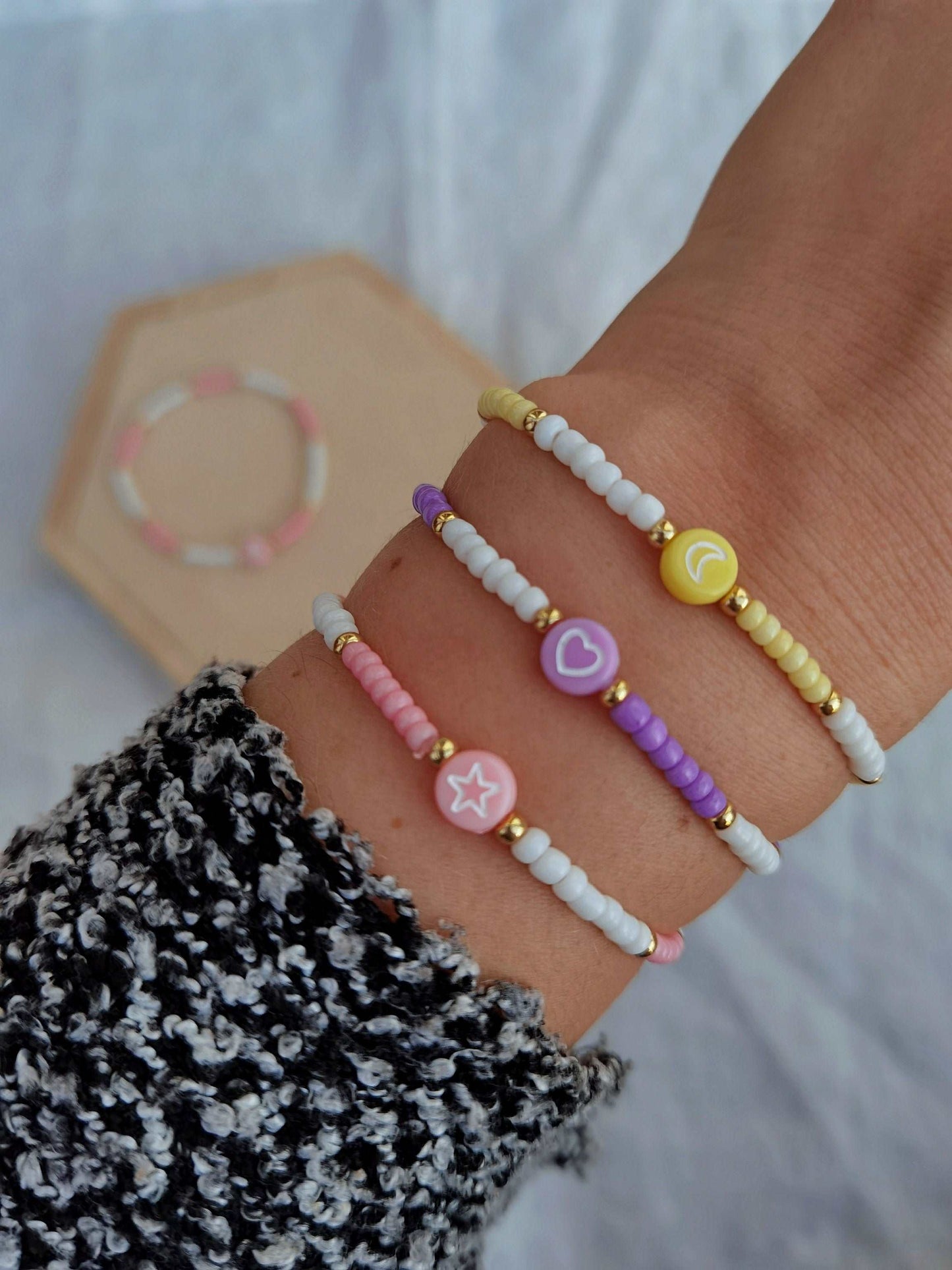 Tragefoto drei bunte Perlenarmbänder. Unteres Armband rosa und weiße Perlen mit einem Stern in der Mitte, mittleres Armband lila und weiße Perlen mit einem Herz in der Mitte und oberes Armand gelbe und weiße Perlen mit einem Mond in der Mitte