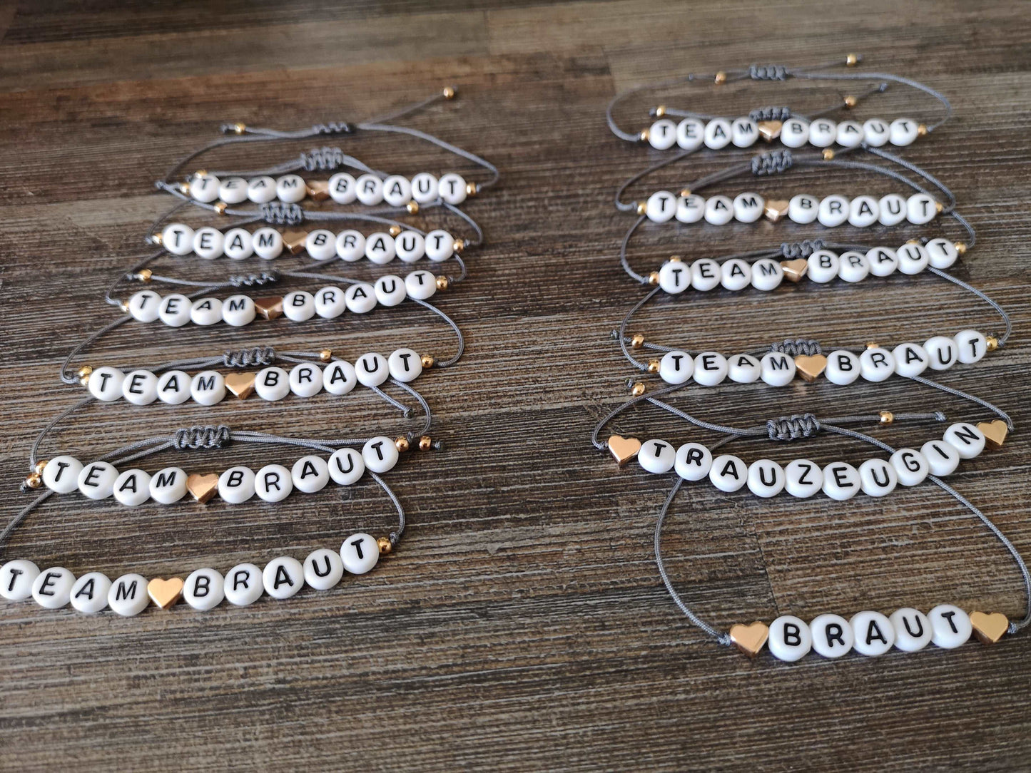 12 personalisierte JGA Armbänder mit grauem Band und roségoldenen Herzen liegen in zwei Reihen aufeinander auf einer dunklen Holzoberfläche