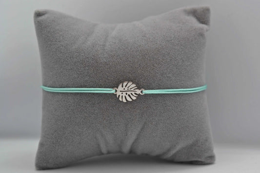 silbernes Monstera Armband mit mintgrünem Band auf einem grauen Schmuckkissen