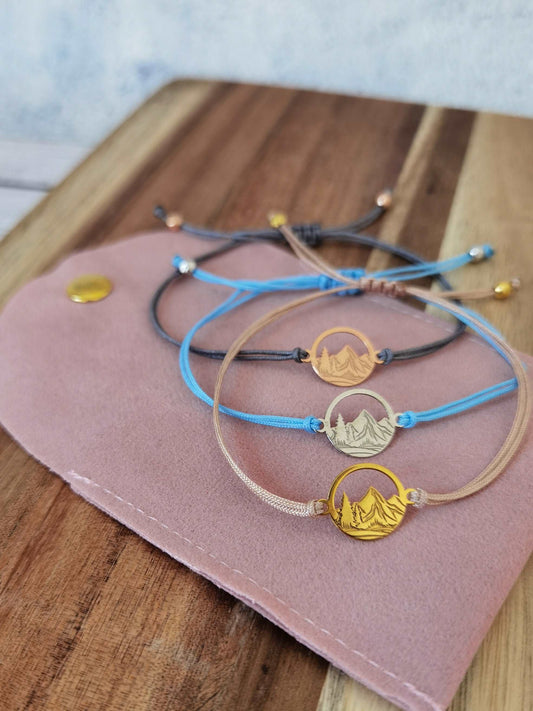 3x Berg Armband als Geschenk für Wanderer liegen auf einer rosa Schmucktasche auf einem dunklen Tisch. Vorderes Armband Beige mit goldenem Berg, mittleres Armband silberner Berg mit babyblauem Band und hinteres Armband grau mit roségoldenem Berg