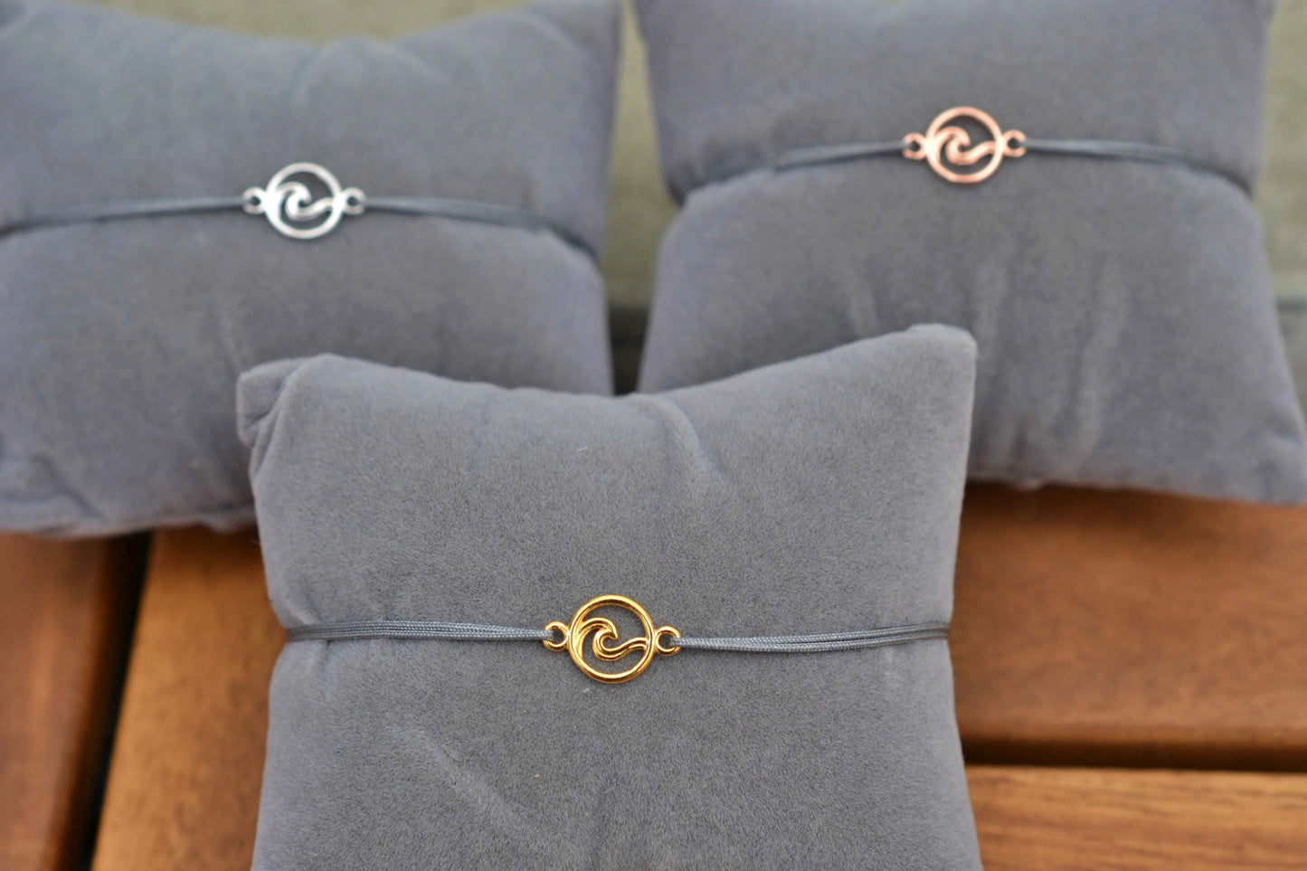 Drei Wellen-Armbänder in gold, silber und roségold jeweils mit grauem Band und auf einem grauen Schmuckkissen