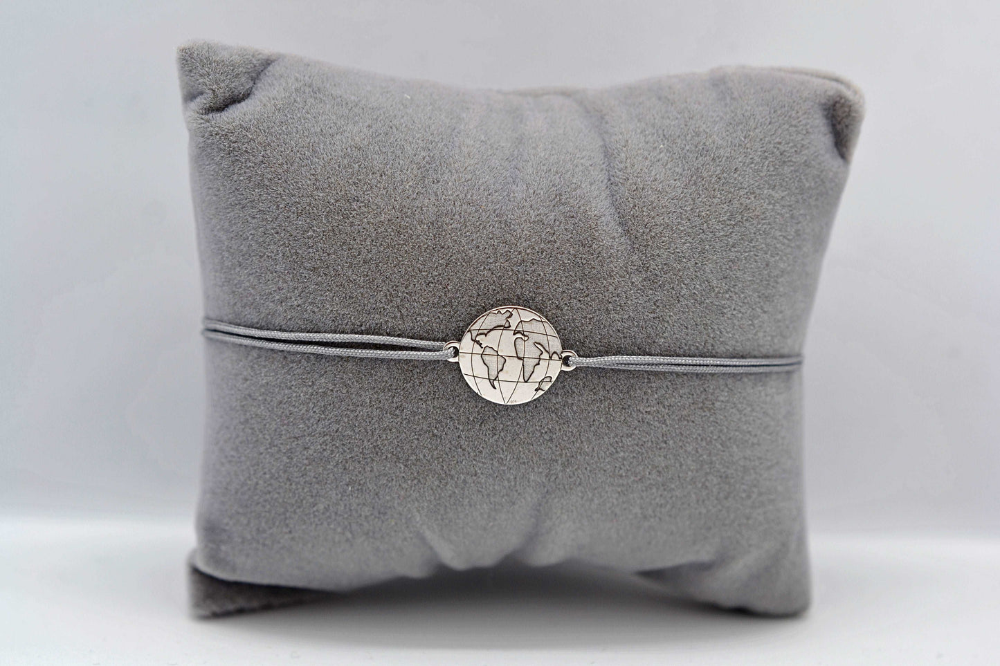 Weltkarte Armband in silber mit grauem Band auf einem grauen Schmuckkissen
