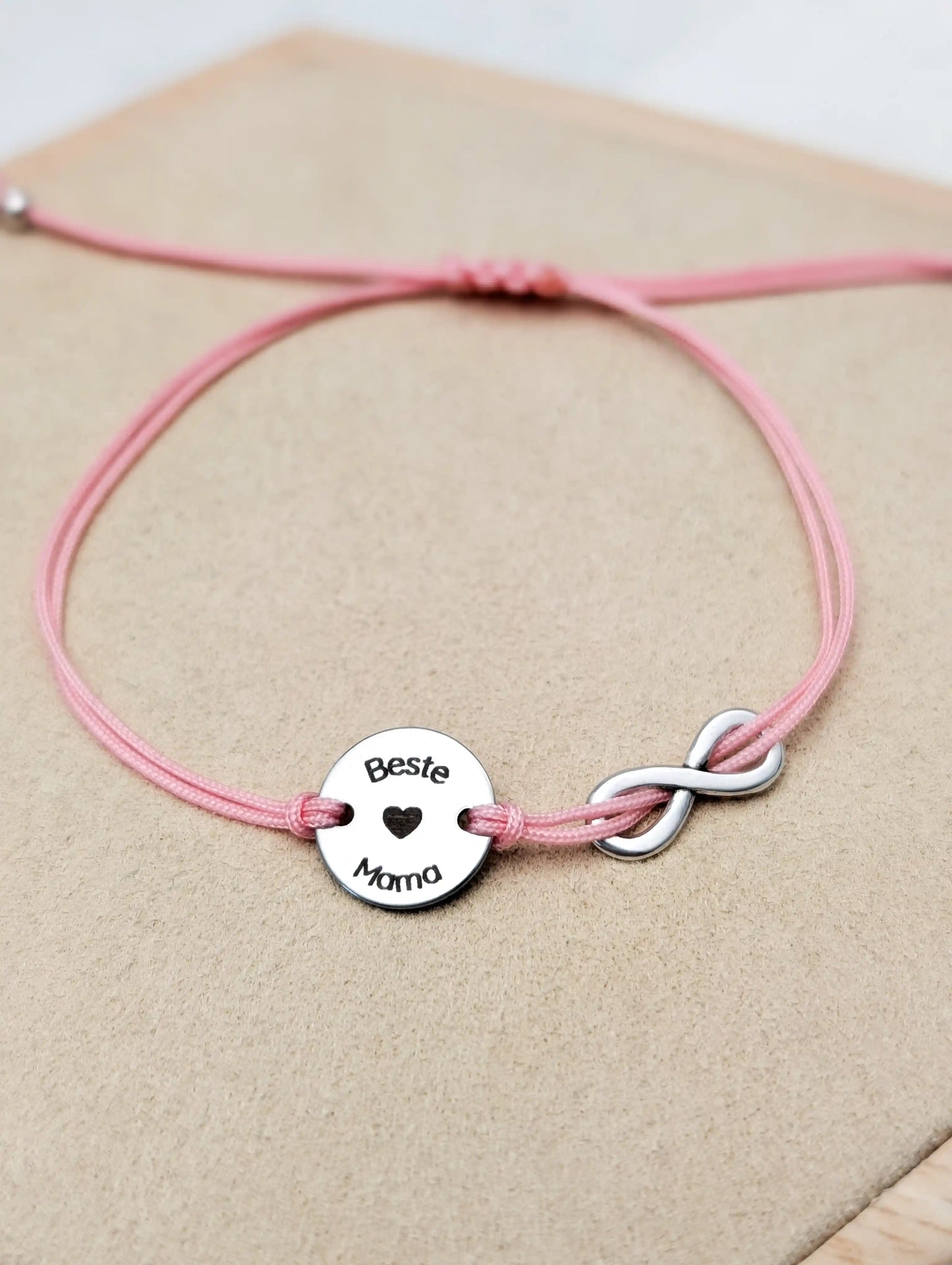 Rosafarbenes Gravur-Armband mit einem silbernen Plättchen mit Gravur "Beste Mama", daneben ein silbernes Unendlichkeitszeichen. Nahaufnahme auf einem Holz Sechseck