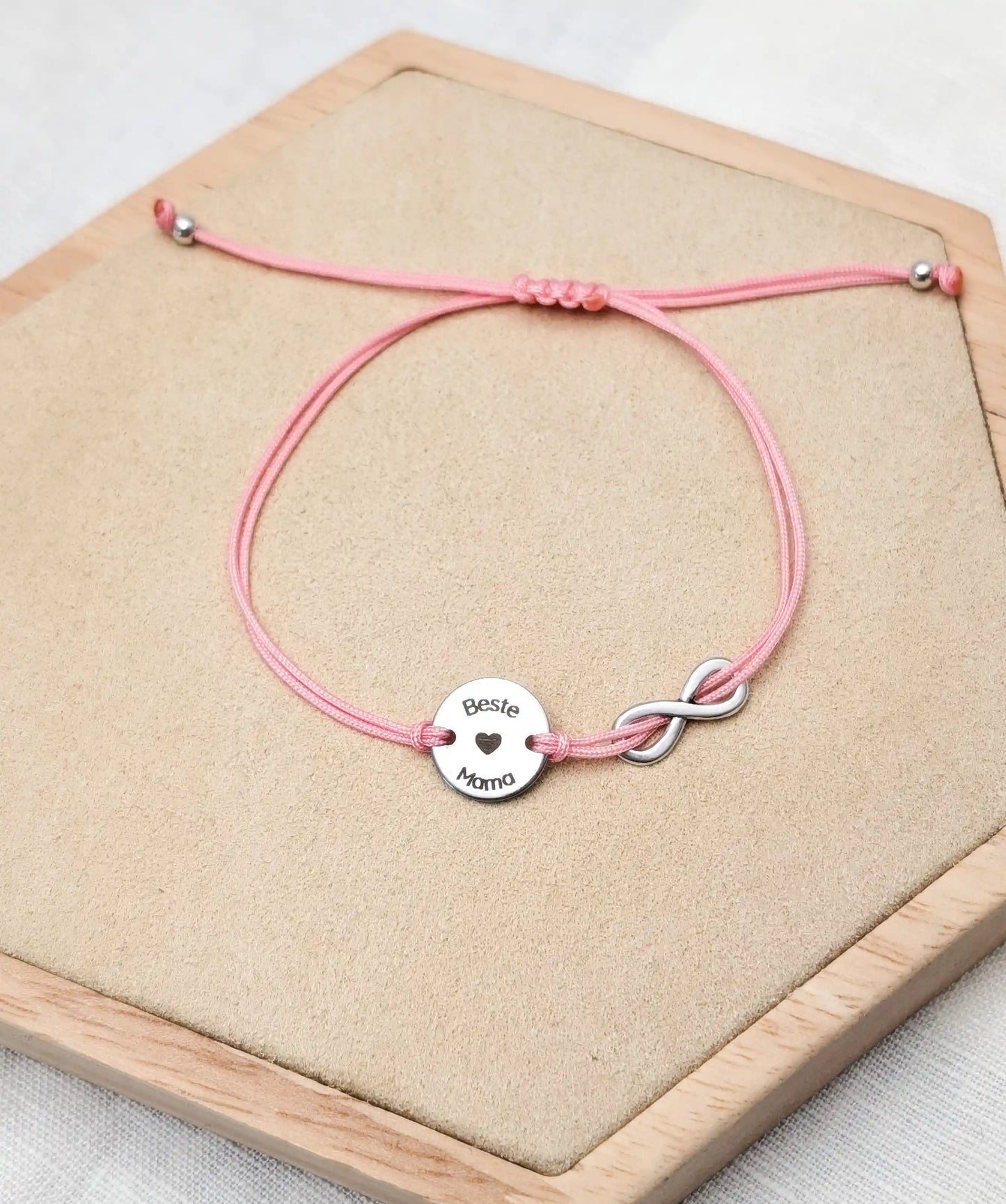 Rosafarbenes Gravur-Armband mit einem silbernen Plättchen mit Gravur "Beste Mama", daneben ein silbernes Unendlichkeitszeichen fotografiert auf einem olz Sechseck