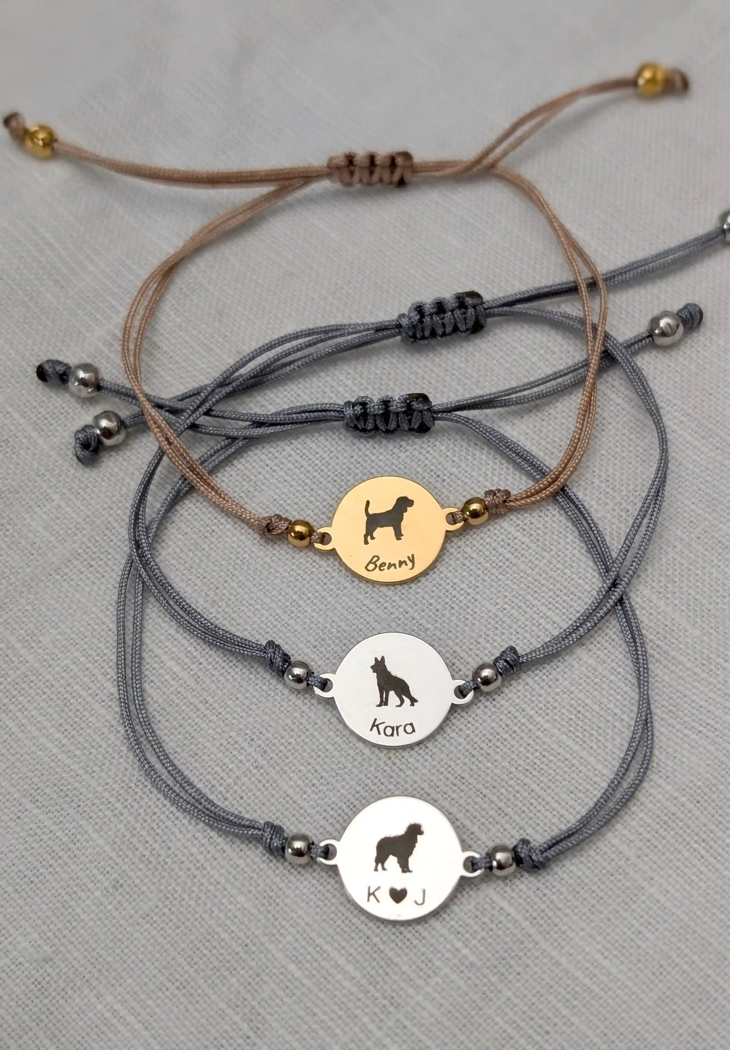 Drei Armbänder mit Hundenamen vor weißem Hintergrund. Graues Armband mit Australian Shepherd und Buchstaben "K herz J", graues Armband mit Schäferhund und "Kara" und beiges Armband mit Beagle und "Benny"