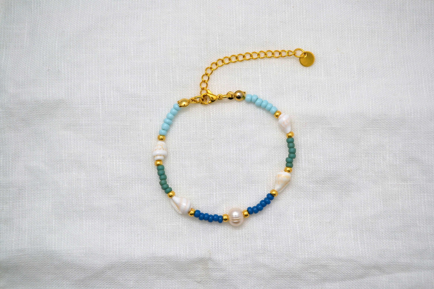 Armband mit vier Muscheln, einer runden Süßwasserperle und blauen, grünen und goldenen Glasperlen vor weißem Hintergrund
