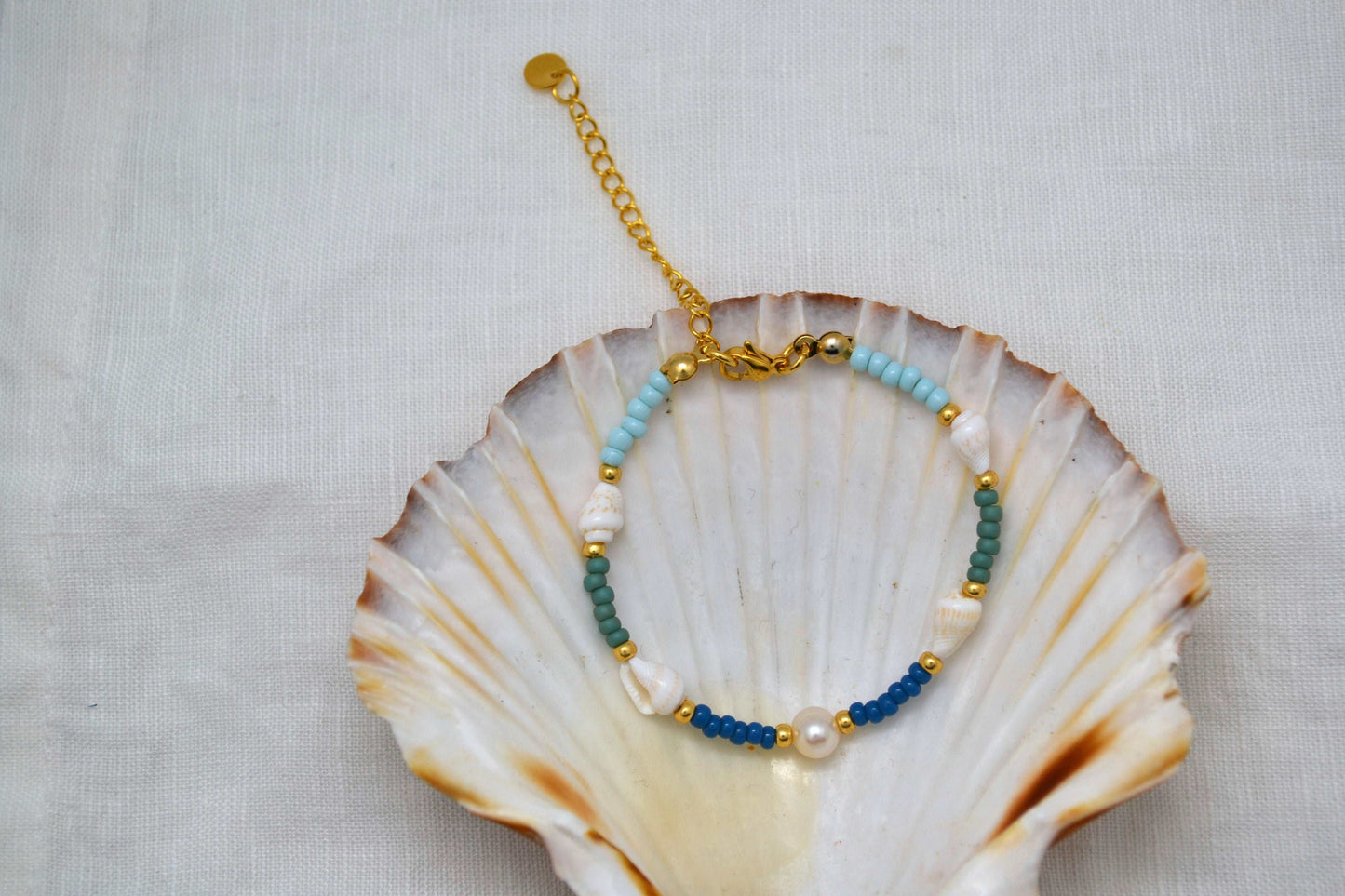 Armband mit Muscheln, einer Süßwasserperle und Glasperlen in blau, grün und gold in einer Muschel dargestellt