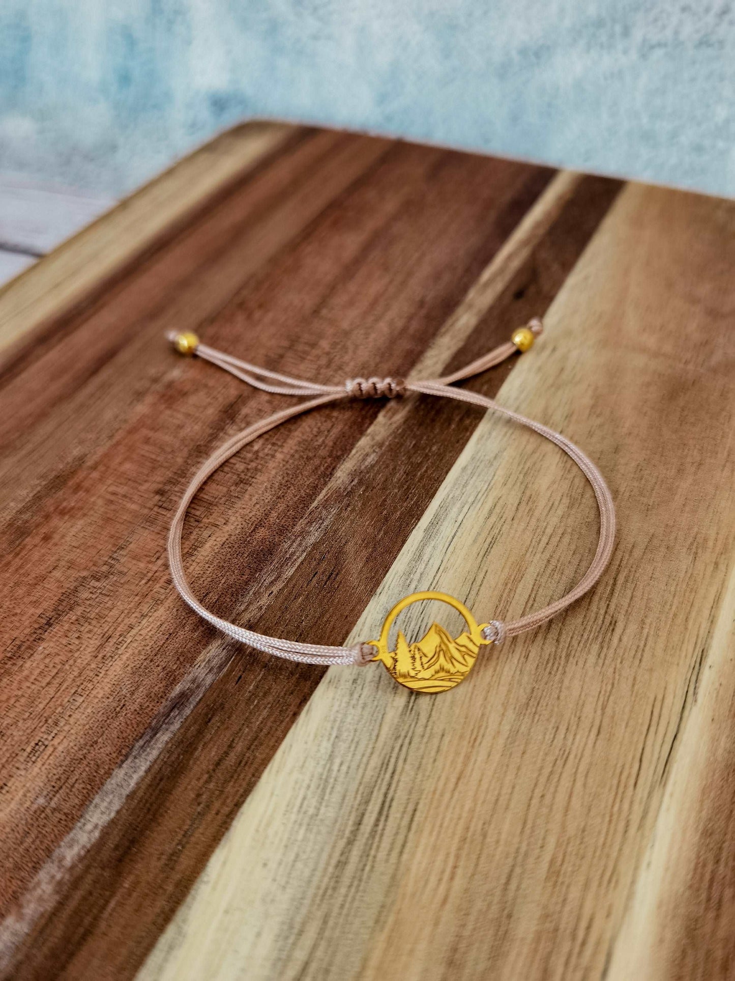 Ein goldenes Berg Armband mit beigem Band liegt auf einem dunklen Holztisch