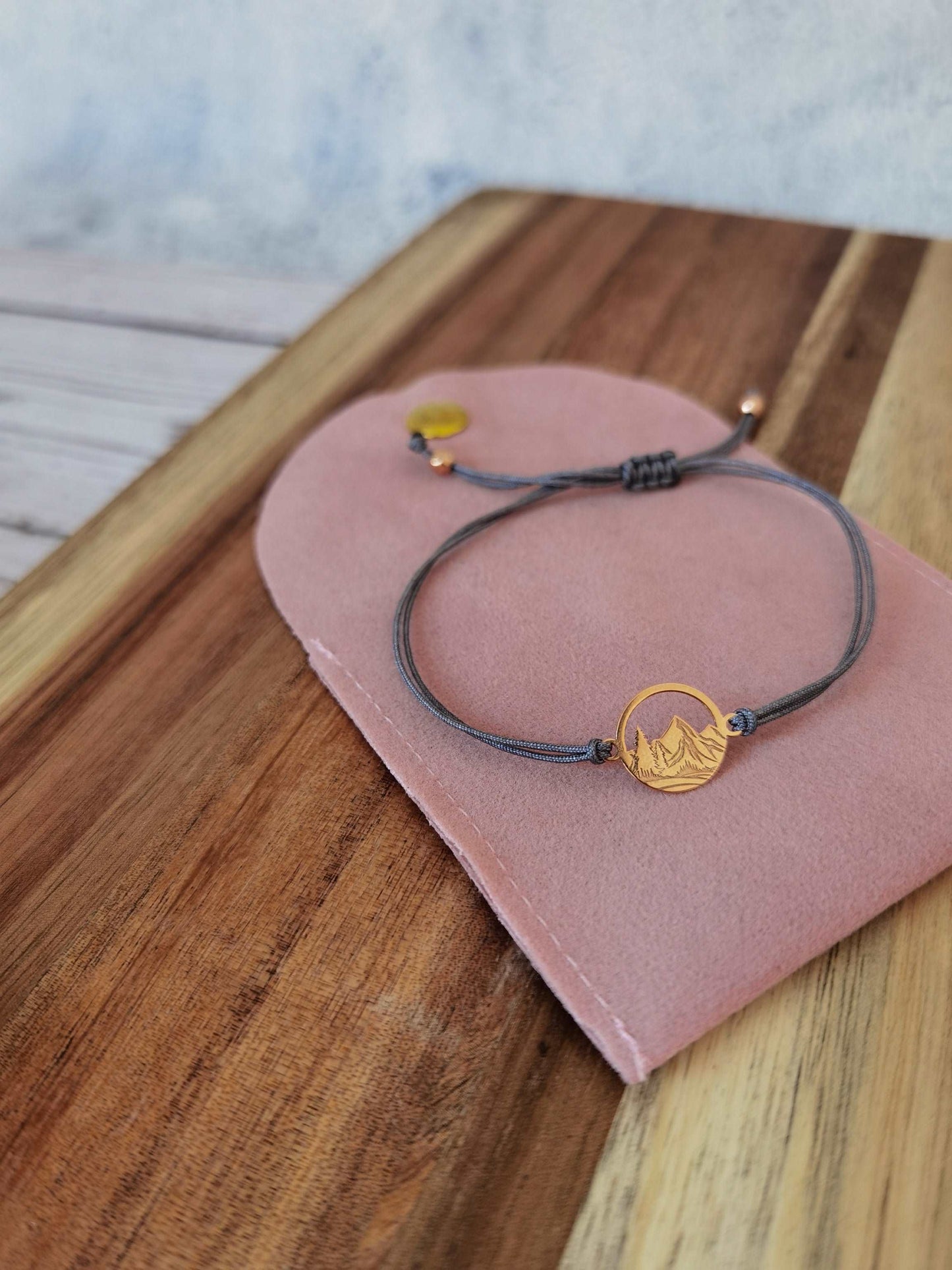 Ein roségoldenes Berg Armband mit grauem Band liegt auf einer rosa Schmucktasche auf einem dunklen Holztisch