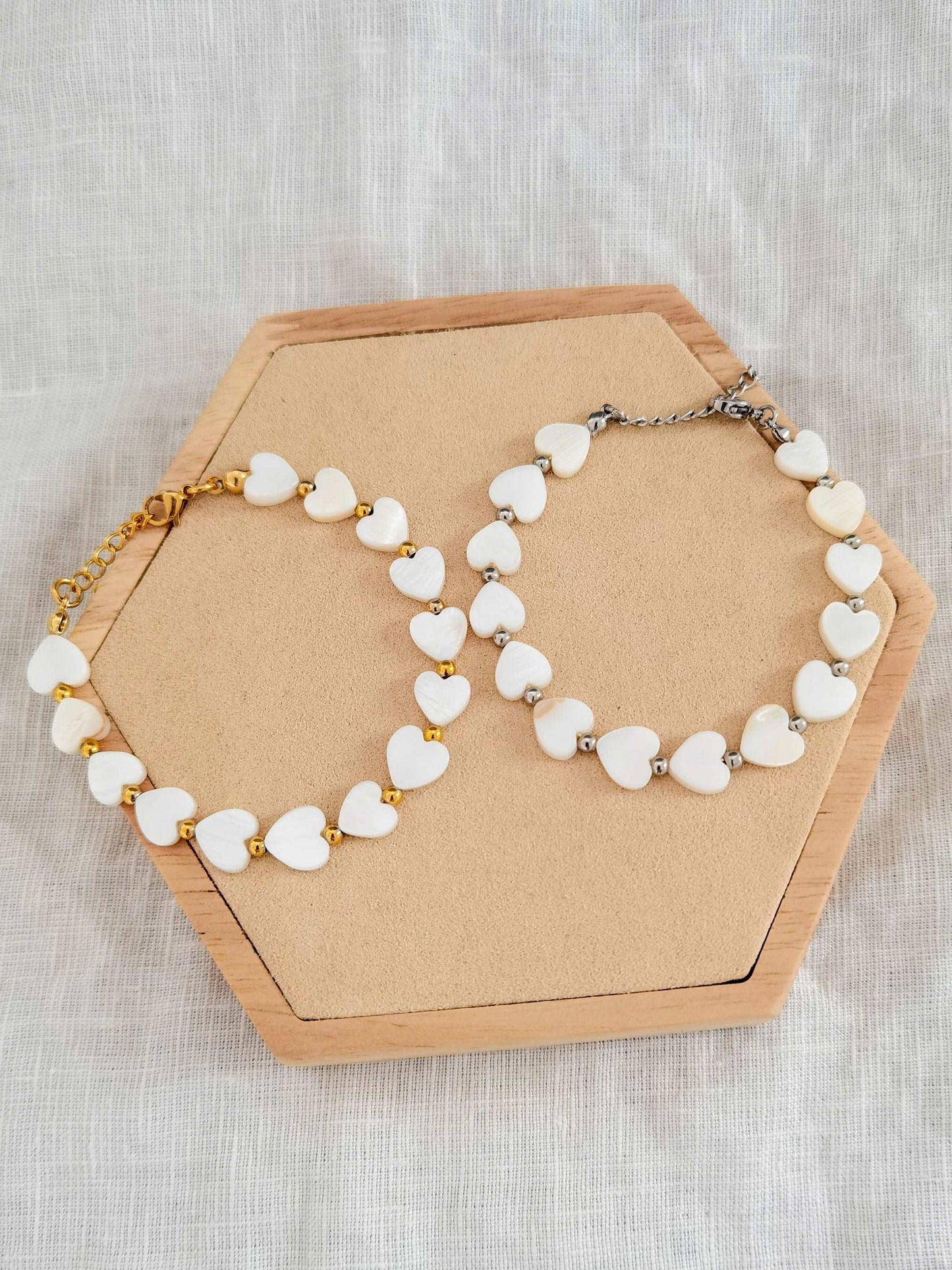 Zwei Herz Perlenarmbänder nebeneinander auf einem Holz Sechseck. Linkes Armband mit goldenen Edelstahl Elementen und linkes Armband mit silbernen