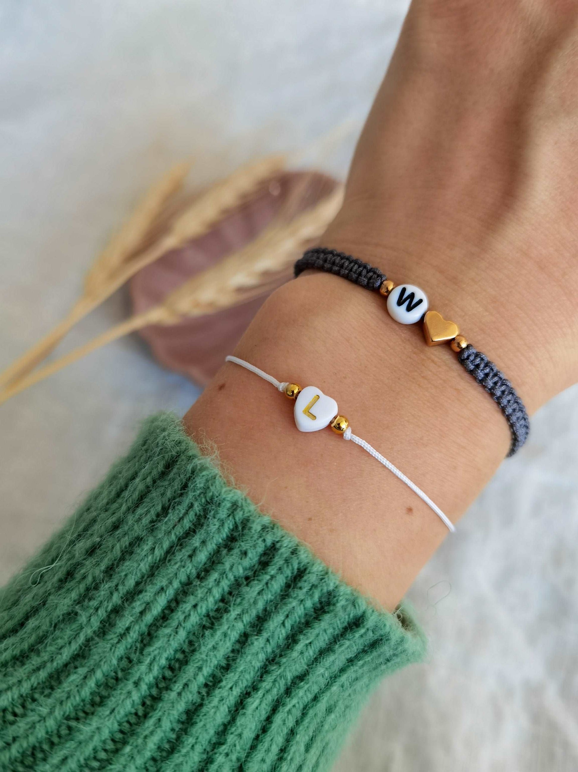 Tragefoto zwei personalisierte Buchstabenarmbänder. Unten weiß goldenes Armband mit Buchstabenherz "L" und oben grau roségoldenes Makramee-Armband mit Buchstabe "W" und einem Herz