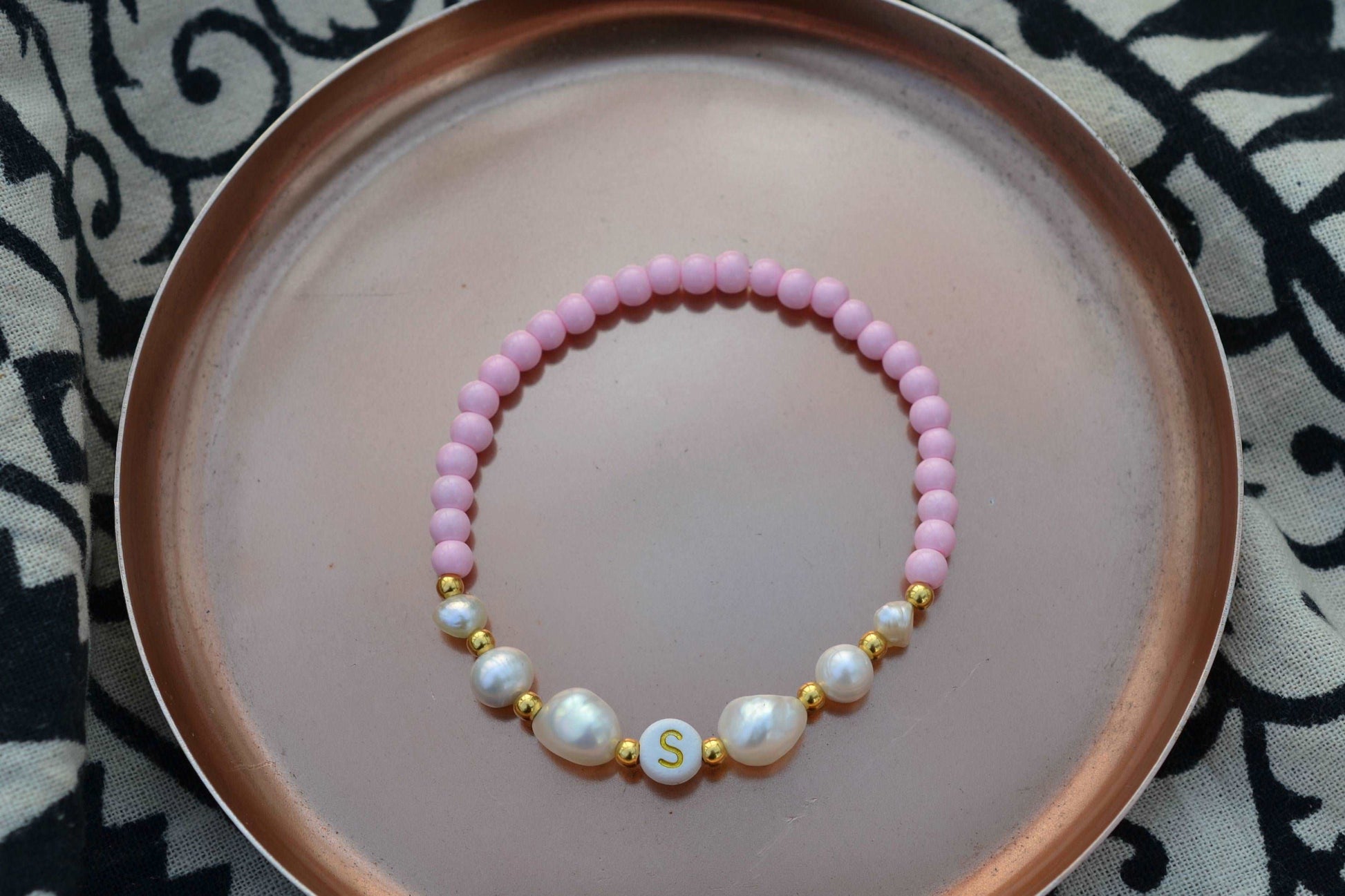 Personalisiertes Süßwasserperlenarmband mit einem Buchstaben nach Wahl und rosa Glasperlen in einer roségoldenen Schale fotografiert