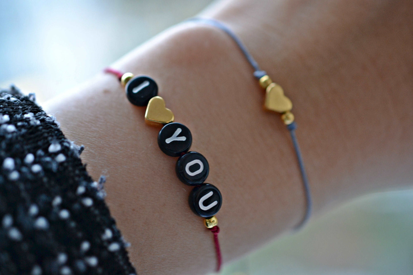 Tragefoto Personalisierbares Buchstabenarmband mit Herz mit rotem Band, schwarzen Buchstabenperlen und goldenem Herz mit der Aufschrift "I" herz "you". Darüber ein zweites Armband nur mit einem goldenen Herz und zwei Perlen sowie einem grauen Band.