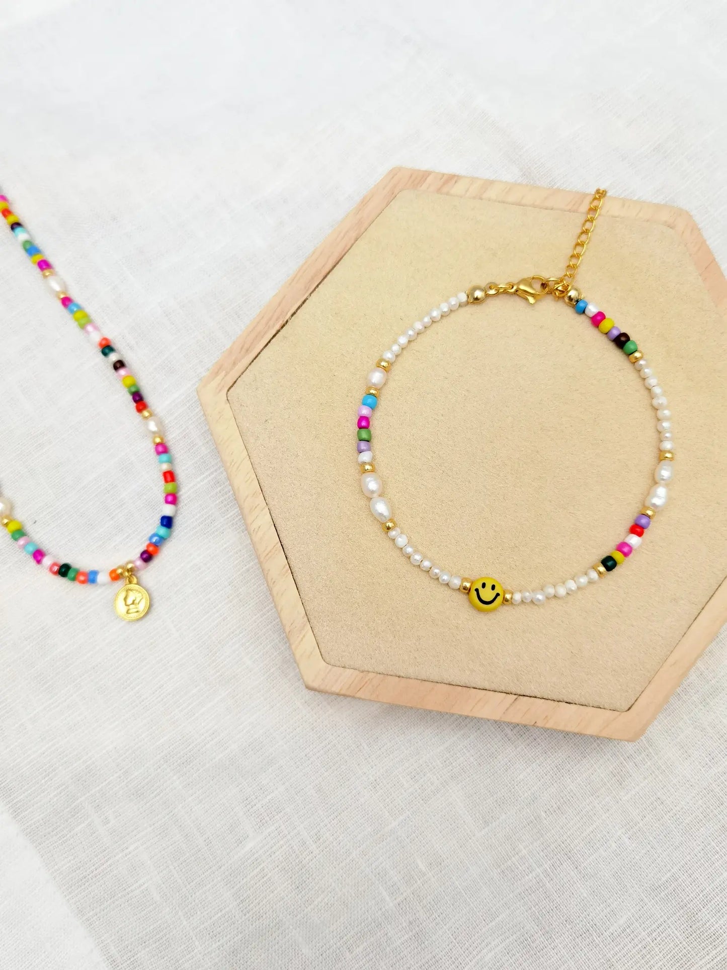Eine Kette aus bunten Perlen und einem goldenen Anhänger in Form einer kleinen Münze und eine Fußkette aus weißen Süßwasserperlen, kleinen bunten Perlen und einem gelben Smiley nebeneinander fotografiert