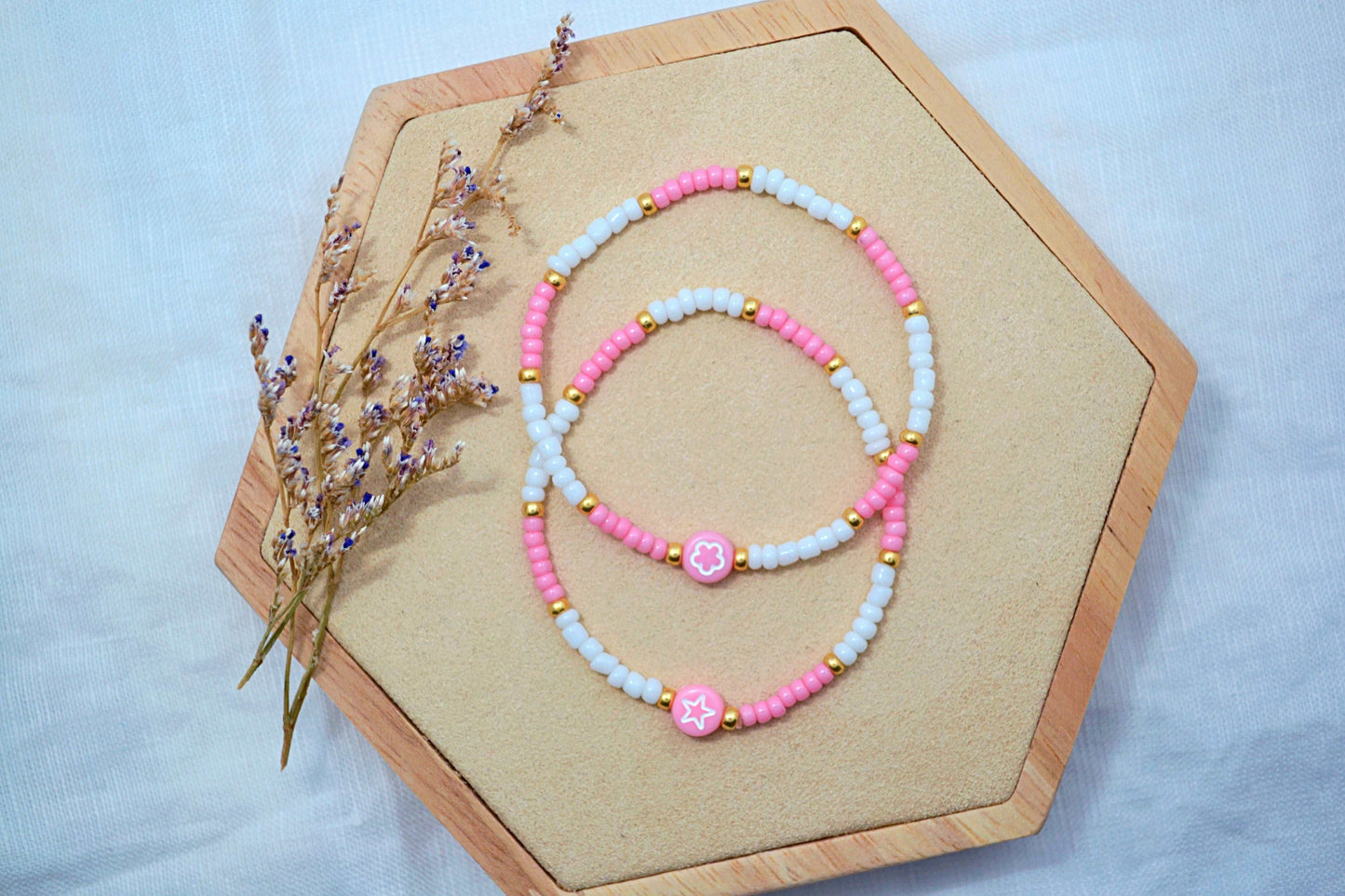 zwei Perlenarmbänder in rosa, weiß und gold mit einer Motivperle (1x Blume, 1x Stern) auf einem Holz Sechseck