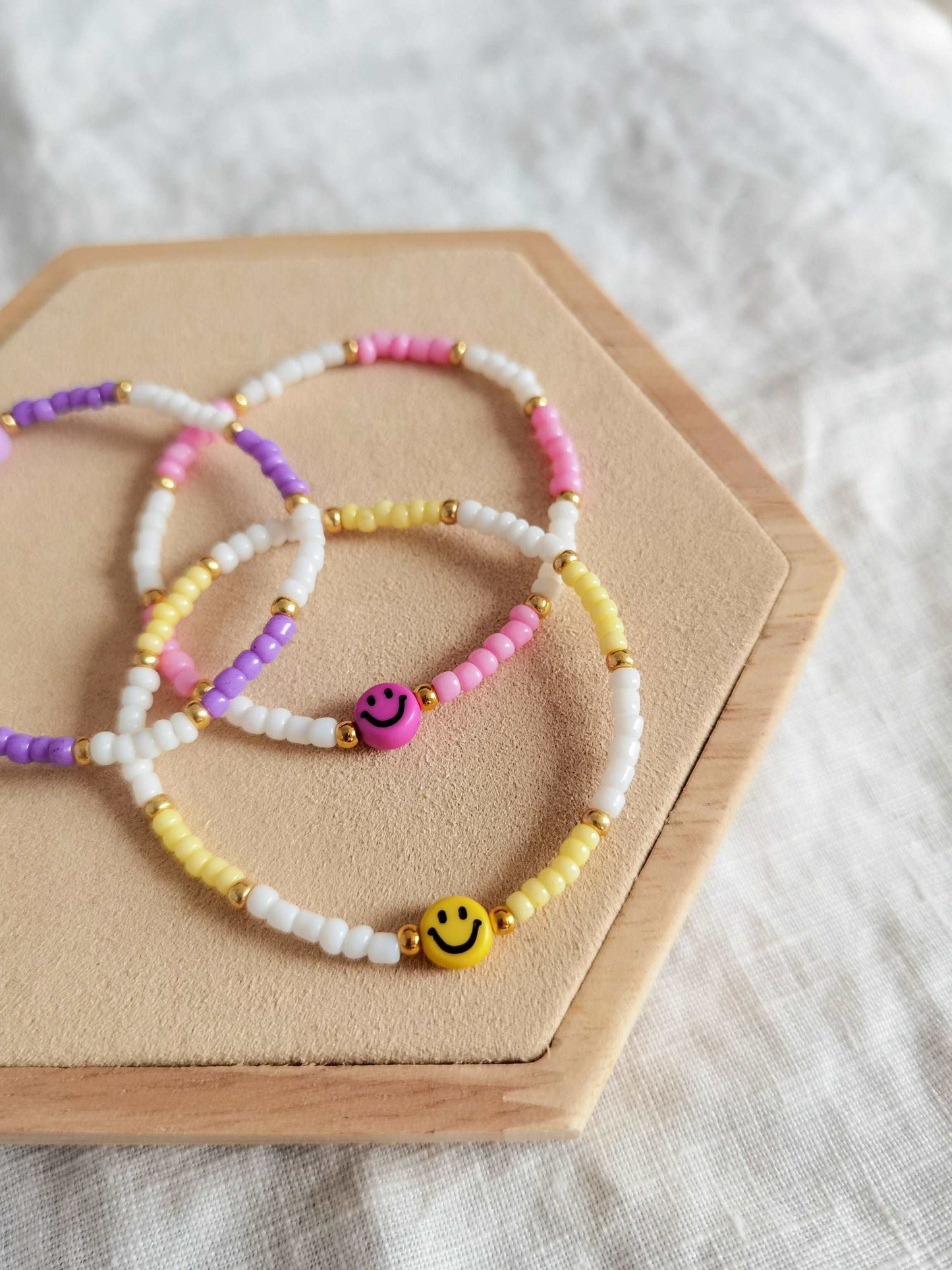 Buntes Smiley Armband in lila, rosa und gelb übereinander gelegt auf einem Holz Sechseck