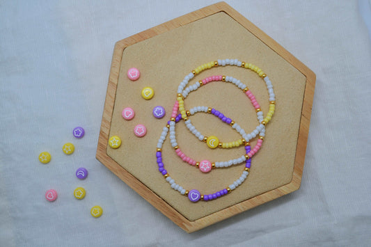 Drei Bunte Perlenarmbänder auf einem Holz Sechseck fotografiert. Daneben diverse bunte Motivperlen