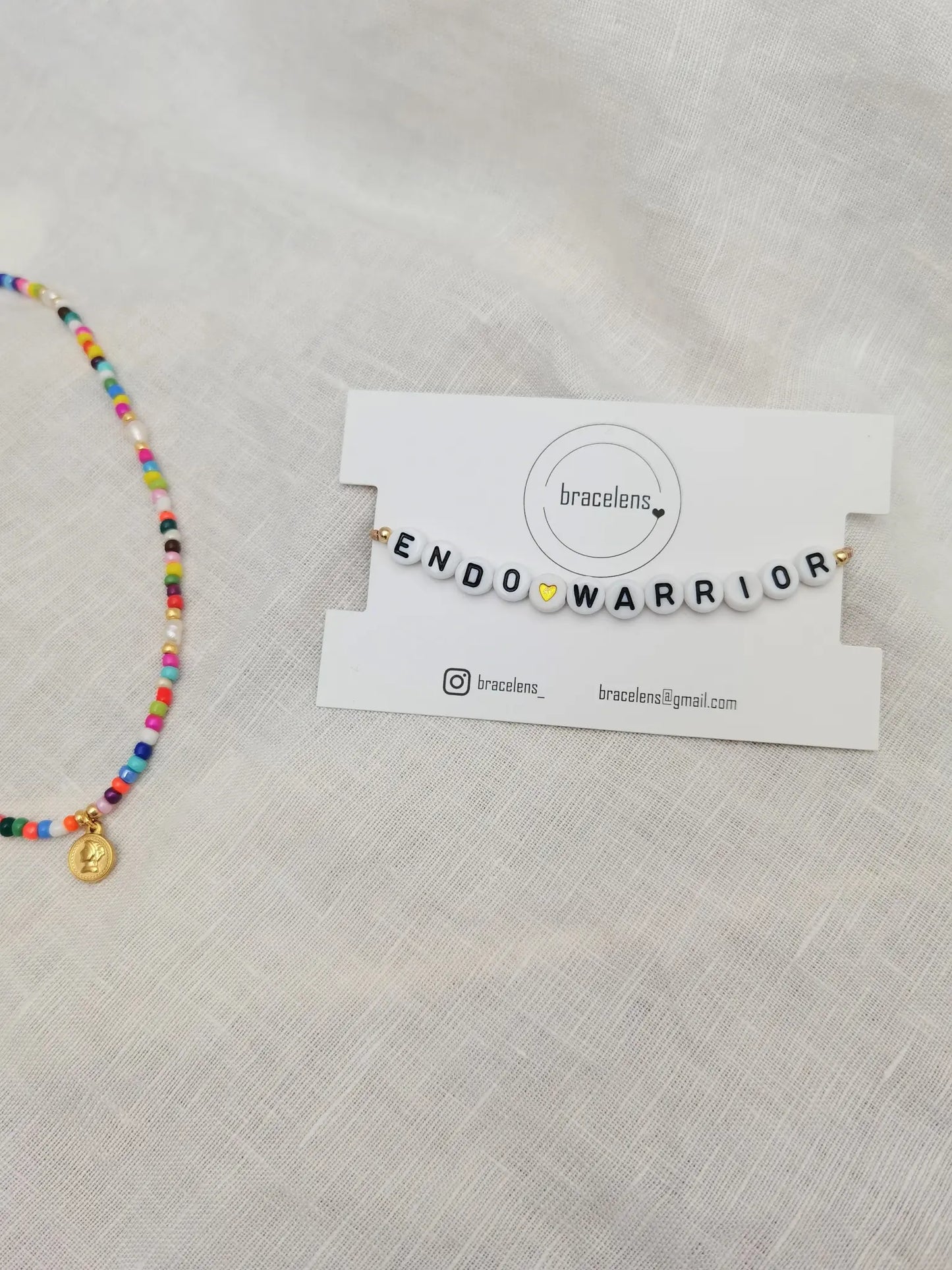 Endo Warrior Armband auf einer weiĂen Schmuckkarte mit bracelens Logo. Daneben eine bunte Perlenkette
