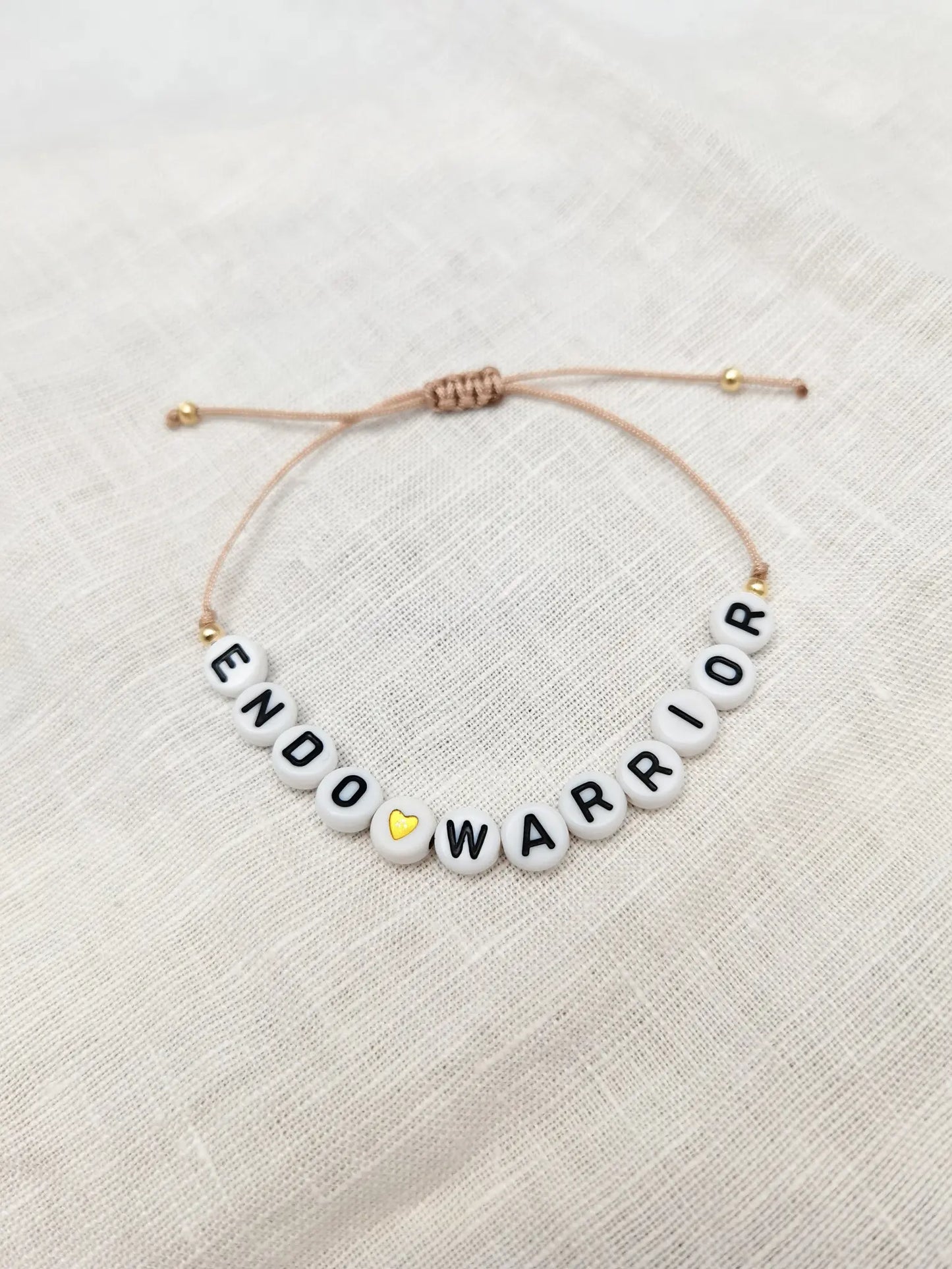 Beiges Endometriose Awareness Armband mit Buchstabenperlen "Endo Warrior" sowie kleinen goldenen Perlen und einem goldenen Herz. Fotografiert auf einem weiĂen Leinentuch
