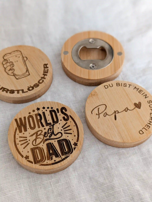 Vier runde Flaschenöffner aus Bambus liegen in zwei Reihen auf einem weißen Tuch. Links oben Flaschenöffner mit Gravur "Durstlöscher", darunter "Papa du bist mein Superheld", daneben "World's best dad" und hinten ein umgedrehter Flaschenöffner, der die magnetische Rückseite zeigt