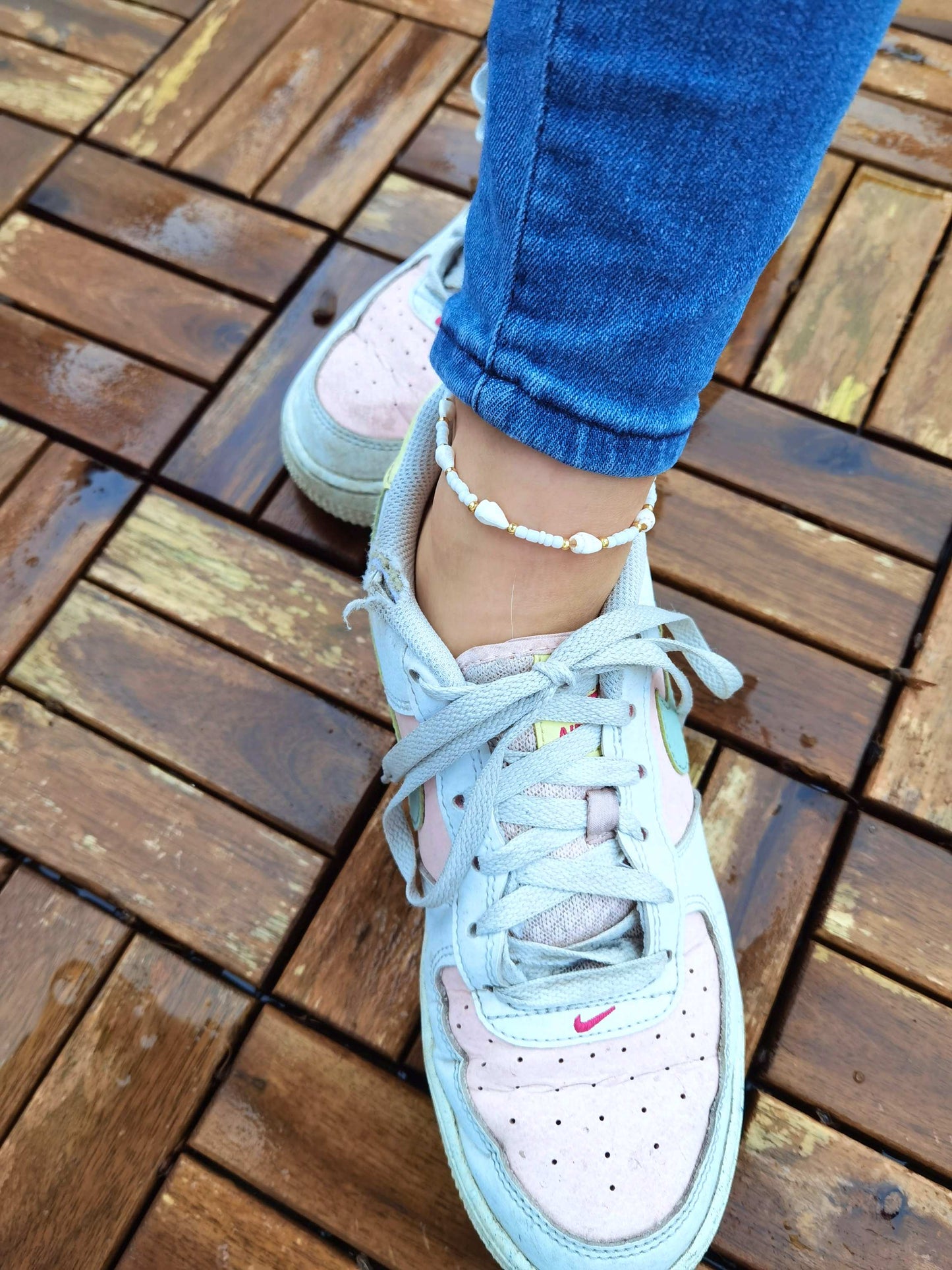 Tragebild einer weiß goldenen Muschel Fußkette am Knöchel einer Frau, die eine blaue Jeans und weiß rosa Nikes trägt