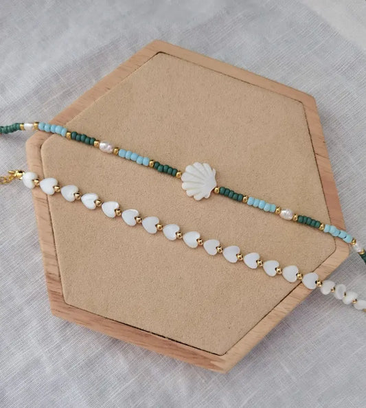 Zwei sommerliche Fußketten auf einem Holz Sechseck. Eine Fußkette mit weißen Herzen und goldenen Perlen und eine Fußkette mit einer weißen Jakobsmuschel und grünen, türkisen und goldenen Glasperlen und weißen Süßwasserperlen