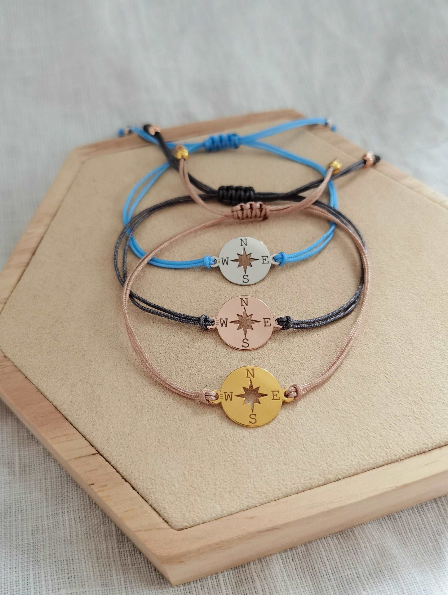 Kompass Armband gold mit beigem Band, roségold mit grauem Band und silber mit hellblauem Band auf einem Holz Sechseck