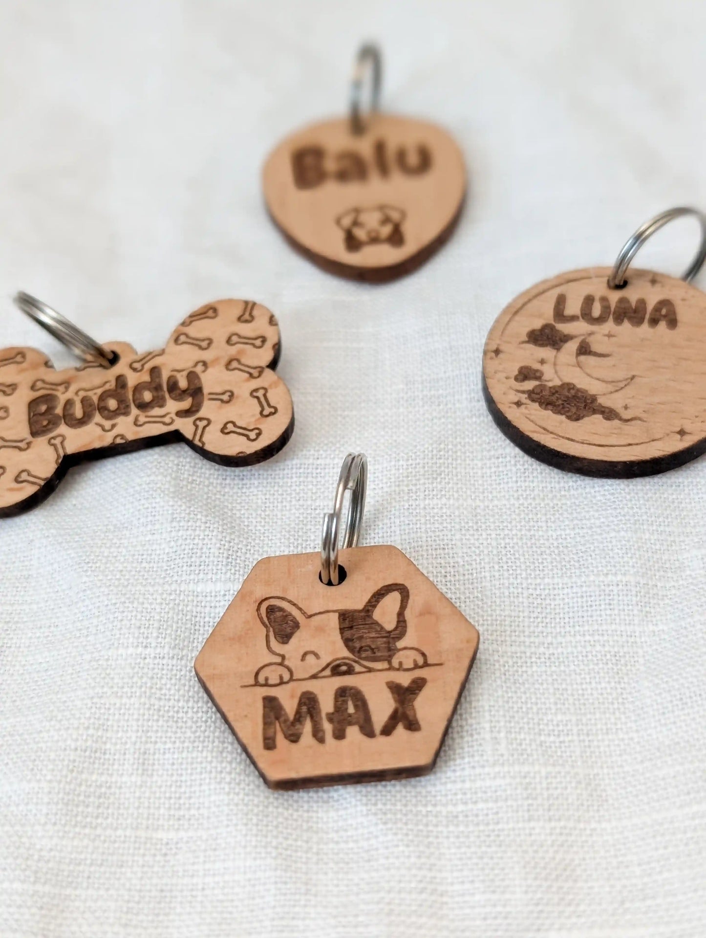Vier personalisierte Hundemarken aus Holz. Vorne ein sechseckiger Anhänger mit einem Hund und "Max", dahinter Knochenform mit Aufschrift "Buddy", daneben ein runder Anhänger mit "Luna" und einem Mond und ganz hinten ein ovaler Anhänger mit "Balu" und einem Hundemotiv