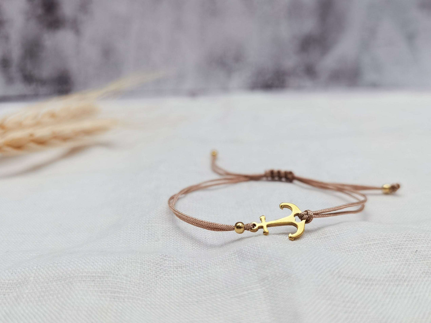 Nahaufnahme goldenes Ankerarmband aus Edelstahl mit beigem Band vor weiß grauem Hintergrund