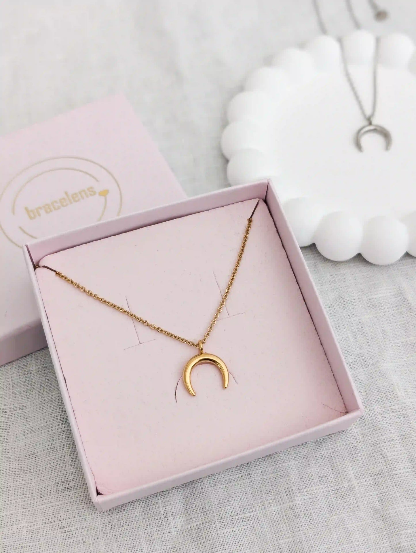 Eine goldene Halbmond Kette in einer rosa Schmuckschachtel. Links im Hintergrund unscharf eine weitere Halbmond Kette in Silber auf einem weiĂźen Gips Untersetzer