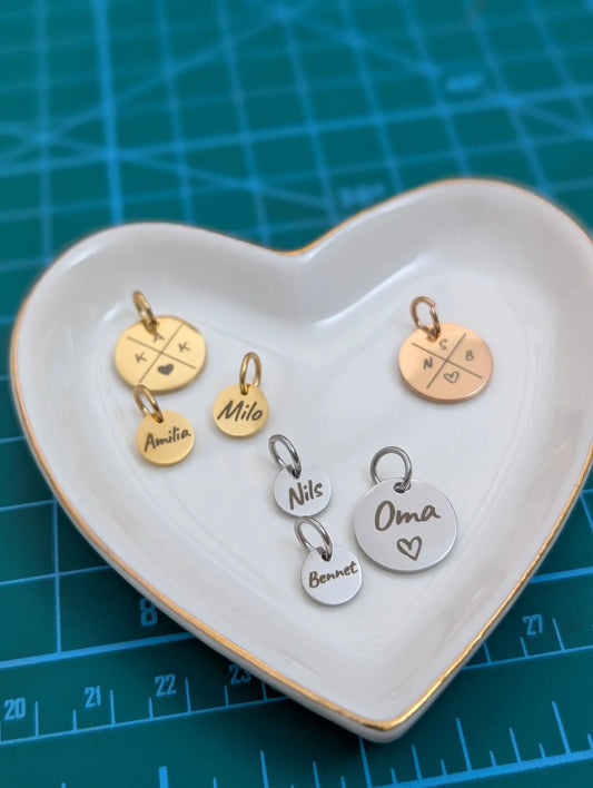 Personalisierte gravierbare Anhänger in Gold und Silber auf einer herzförmigen Schmuckschale – Familienanhänger mit Initialen, Namen und Herzsymbolen als Erweiterung für Namensketten.