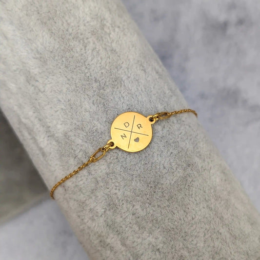 Goldenes Gravurarmband mit drei Buchstaben und einem Herz Symbol in einem Kreuz angeordnet, fotografiert auf grauem Samt