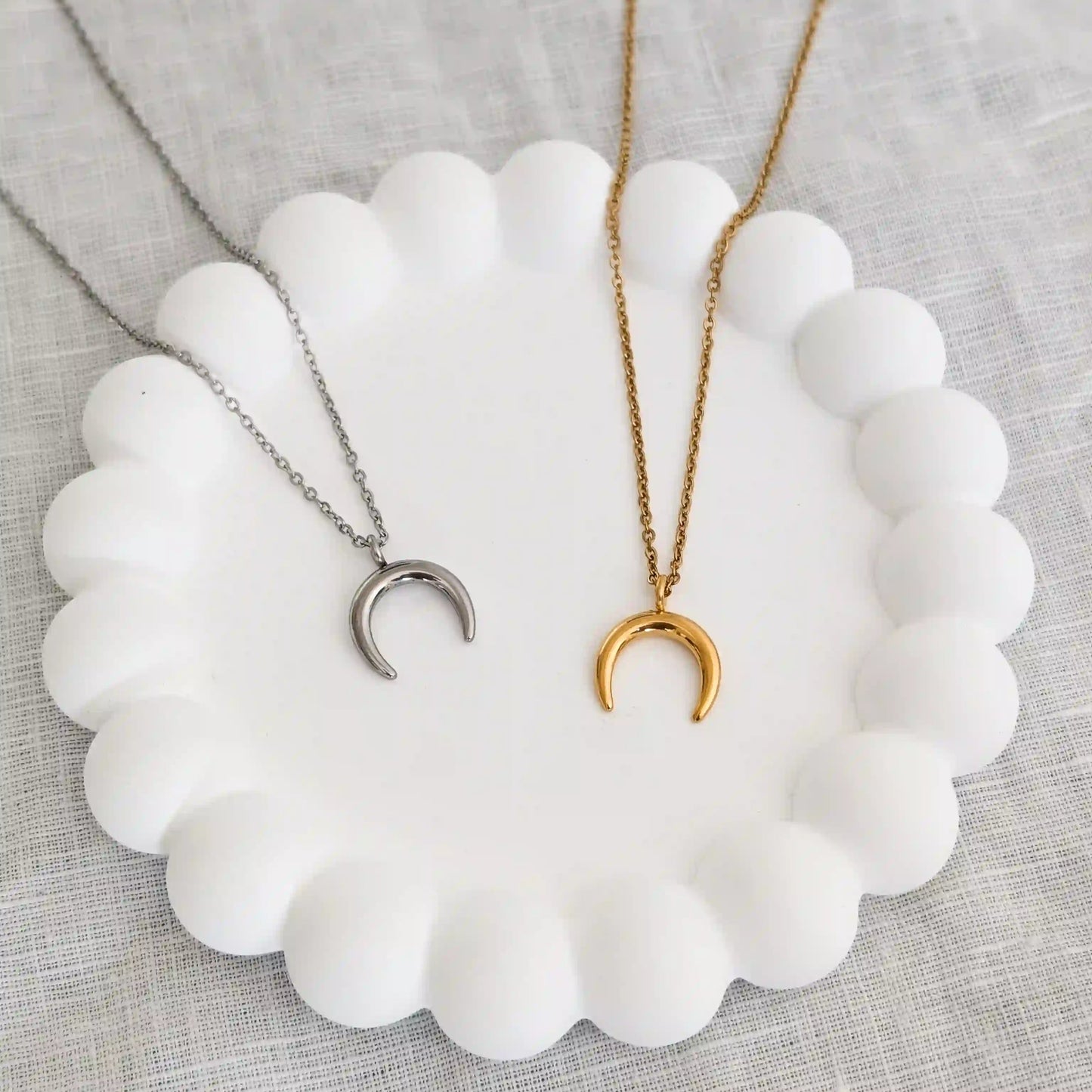 Zwei Halbmond Ketten aus Edelstahl liegen nebeneinander auf einem weiĂźen Gips Untersetzer. Links Halbmond Kette in silber und rechts in gold.