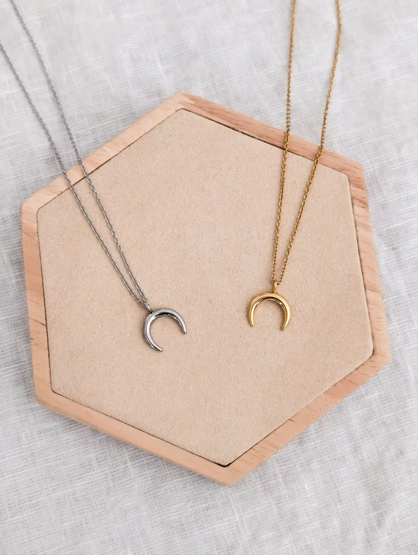 Zwei Halbmond Ketten aus Edelstahl liegen nebeneinander auf einem Holz Sechseck. Links Halbmond Kette in silber und rechts in gold.
