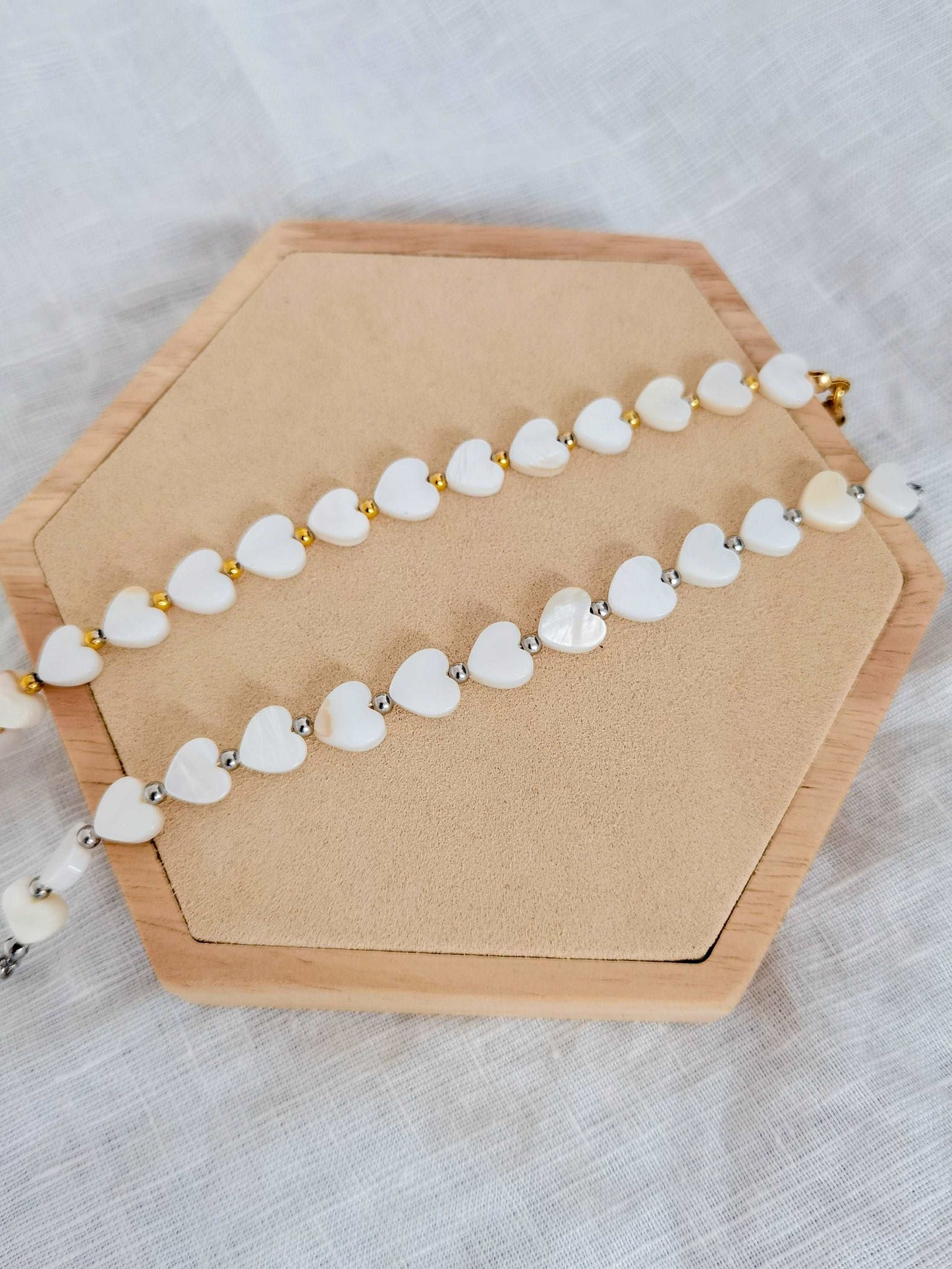 Zwei Herz Perlenarmbänder untereinander auf einem Holz Sechseck. Oberes Armband weiß und gold und unteres weiß und silber