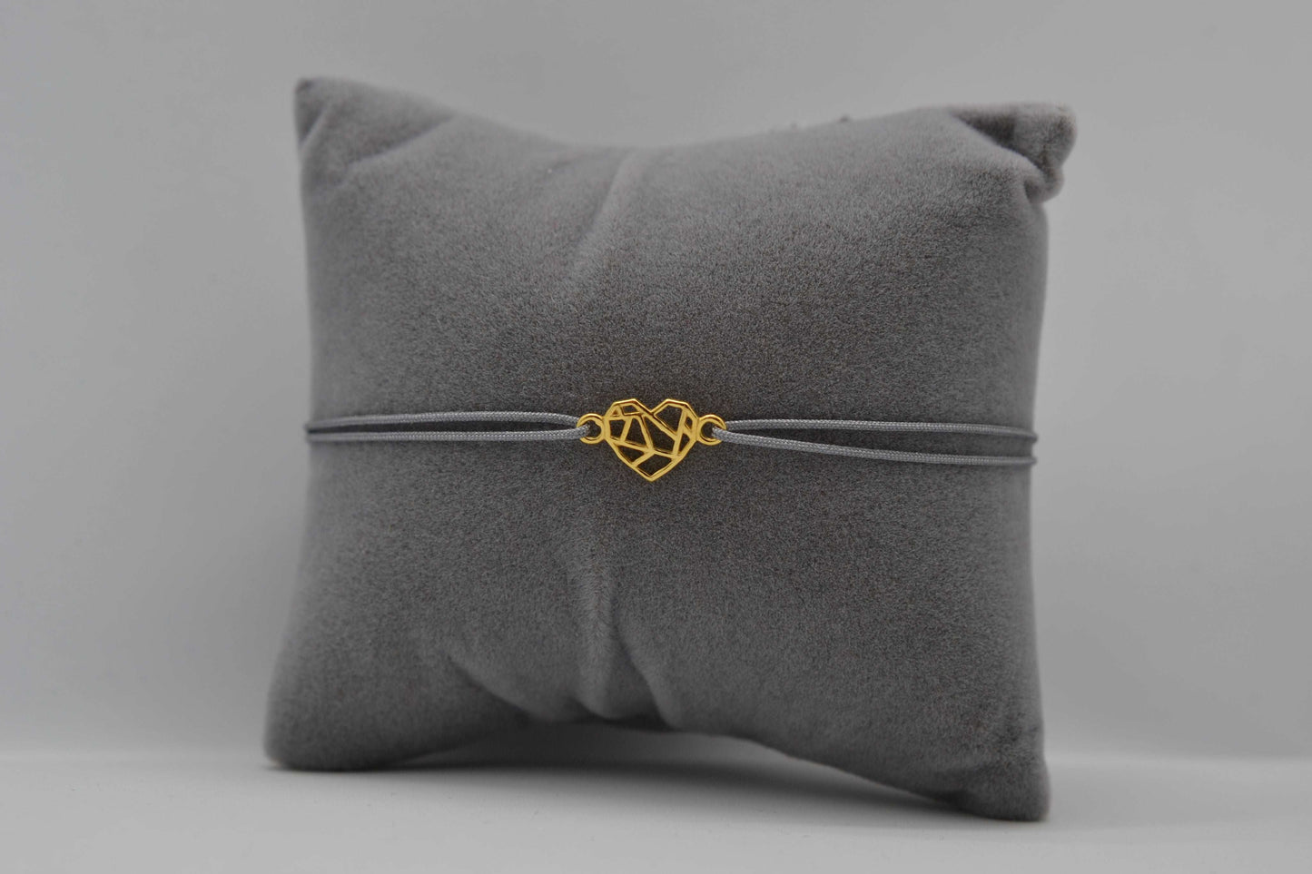 Goldenes Origami Herz Armband mit grauem Band auf einem grauen Schmuckkissen
