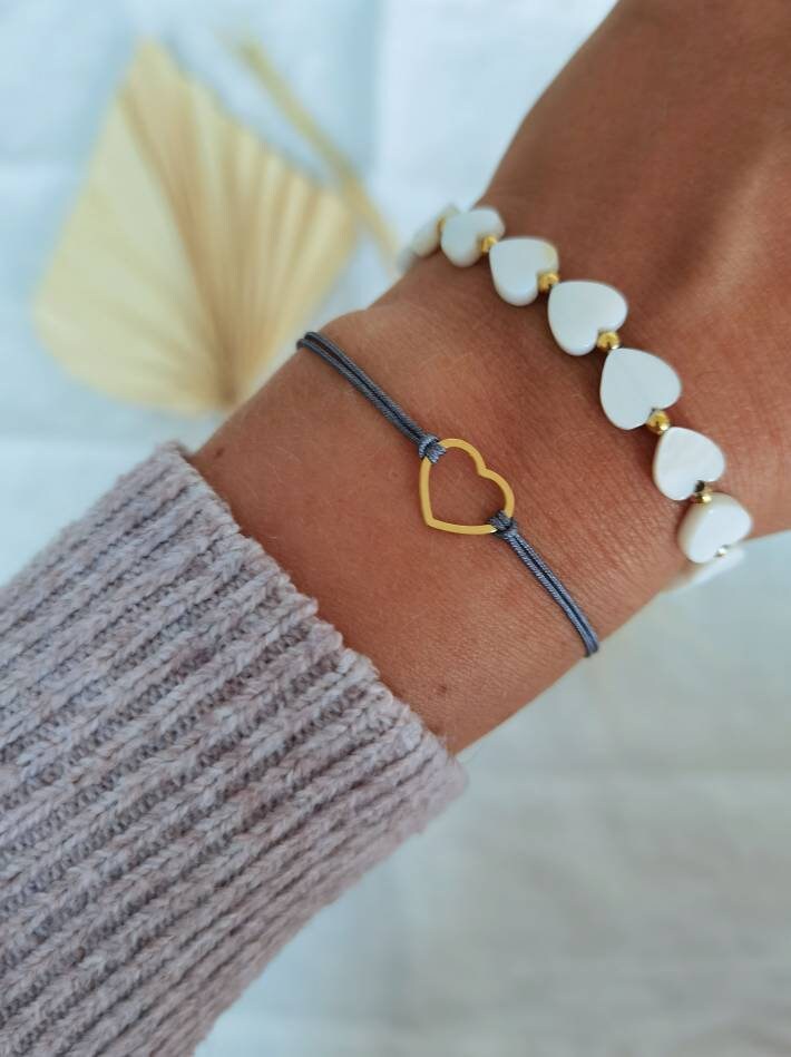 Tragefoto zwei Herz Armbänder. Oberes Armband aus weißen Muschelperlen in Herzform und kleinen goldenen Perlen und unteres Armband aus einem goldenen Herz und grauem Band