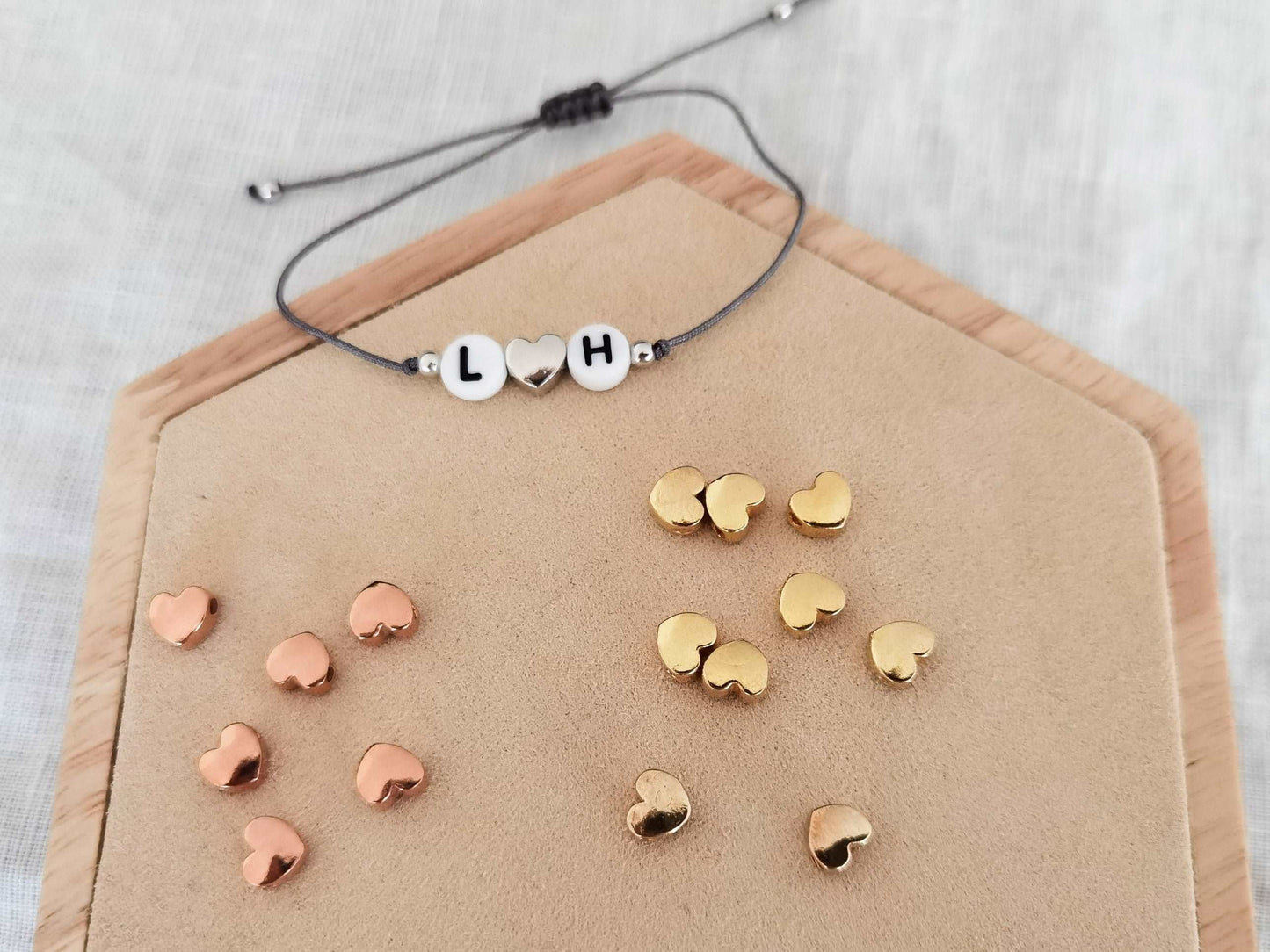 Ein personalisierbares Initialen Armband mit einem silbernen Herz liegt auf einem Holz Sechseck. Vor dem Armband liegen mehrere Herzperlen in roségold und gold