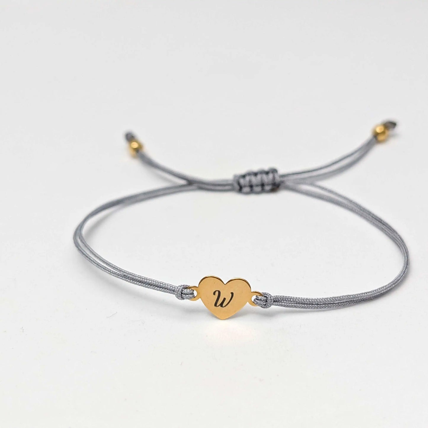 Graues Armband mit einem goldenen Herz, das mit dem Buchstaben "W" graviert ist, vor weißem Hintergrund