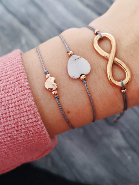 Tragefoto drei Armbänder 1x Armband mit Herzperle in roségold, 1x Armband Muschelherz mit roségoldenen Perlen, 1x Armband Unendlichkeitszeichen in roségold. Alle mit grauem Band