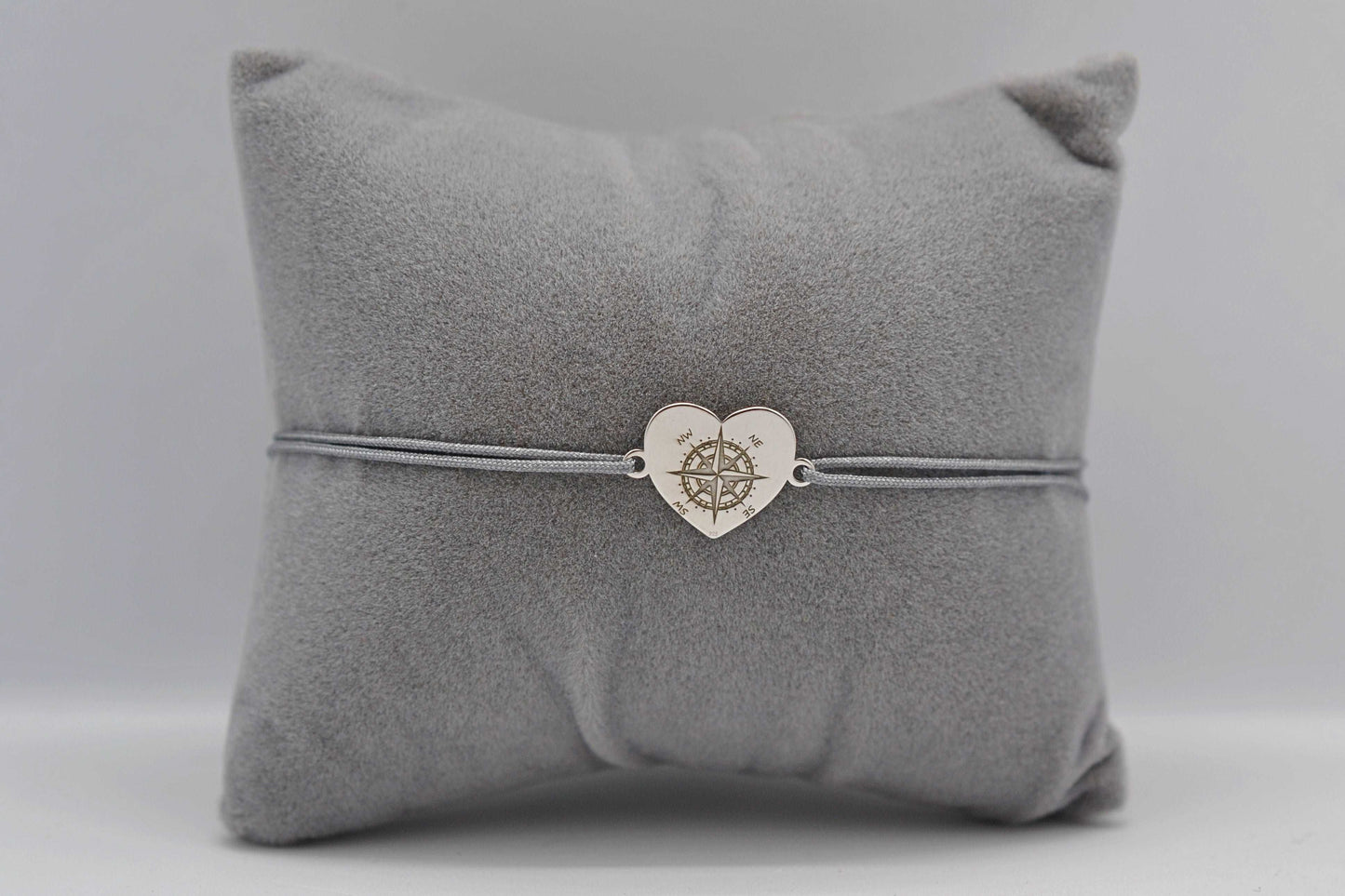 Herz Kompass Armband für Reiseliebhaber mit silbernem Herzen und grauem Band fotografiert auf einem grauen Schmuckkissen