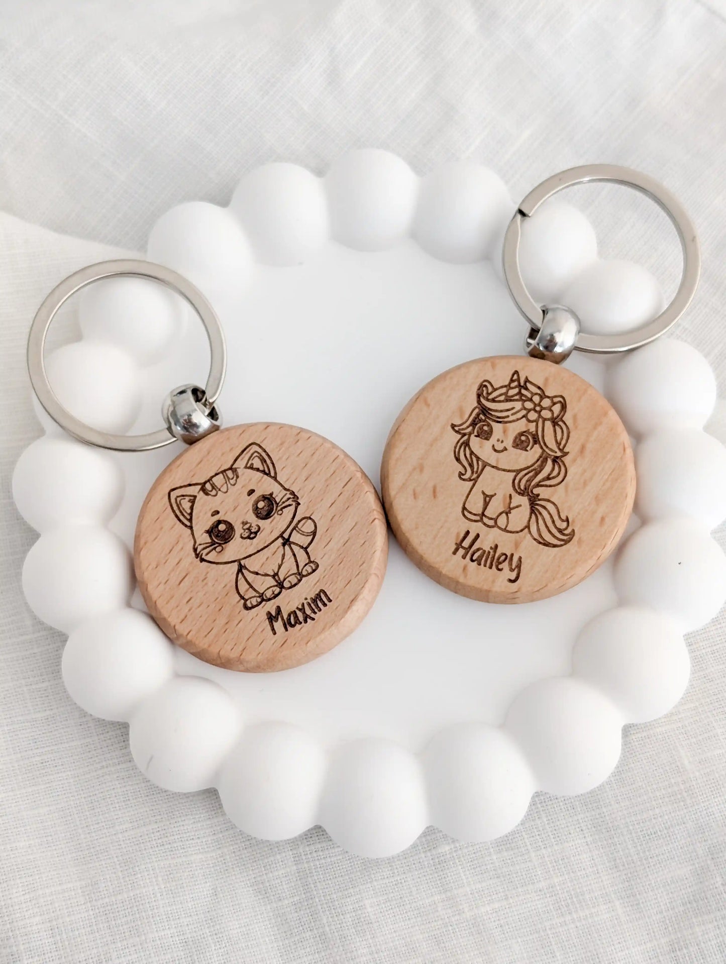 Zwei personalisierte Schlüsselanhänger aus Holz für Kinder liegen nebeneinander in einer weißen Schmuckschale. Links ein Anhänger mit einer Katze und dem Namen Maxim und rechts ein Anhänger mit einem Einhorn und dem Namen Hailey
