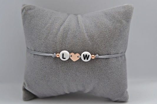 Initialen Armband mit rosévergoldetem 925 Sterling Silber Herz, grauem Band und den Buchstaben L und W. Fotografiert auf einem grauen Schmuckkissen.