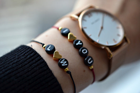 Zwei personalisierbare Armbänder mit schwarzen Buchstabenperlen und goldenen Herzen an dem Handgelenk einer Frau. Ein Armband mit Aufschrift "L" herz "W" und eins mit "I" herz "you"