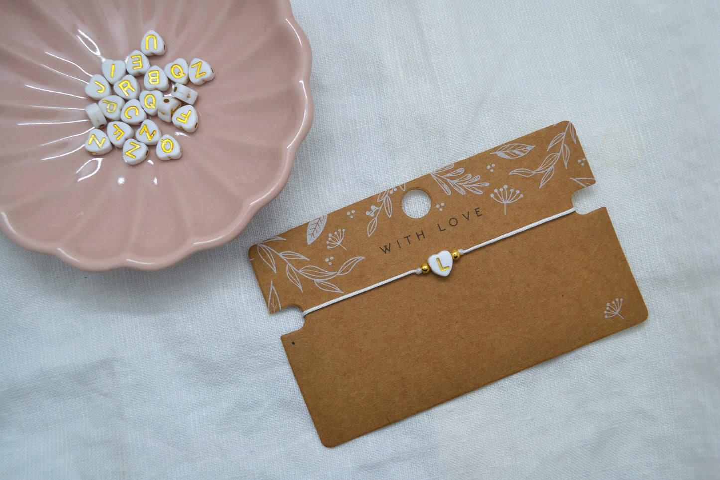 Personalisiertes Armband mit Herzbuchstabe "L" mit goldenem Buchstaben und weißem Band vor einer rosa Schmuckschale mit weiteren Buchstabenperlen