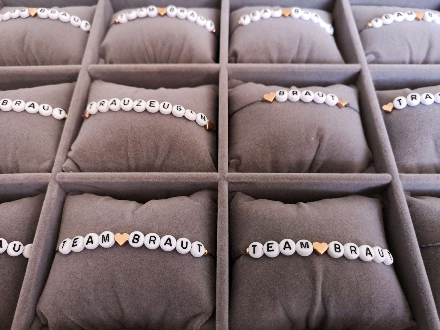 12 personalisierte JGA Armbänder mit grauem Band und roségoldenen Herzen nebeneinander auf grauen Schmuckkissen