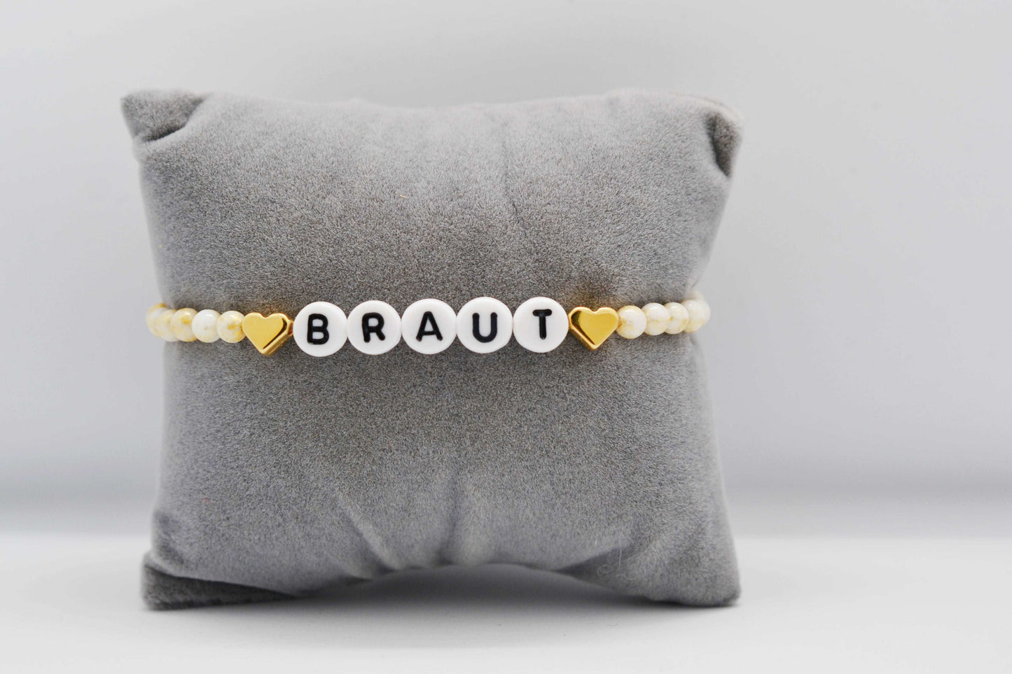 Personalisierbares JGA Perlenarmband mit goldenen Herzen und gold weißen Jadeperlen auf einem grauen Schmuckkissen