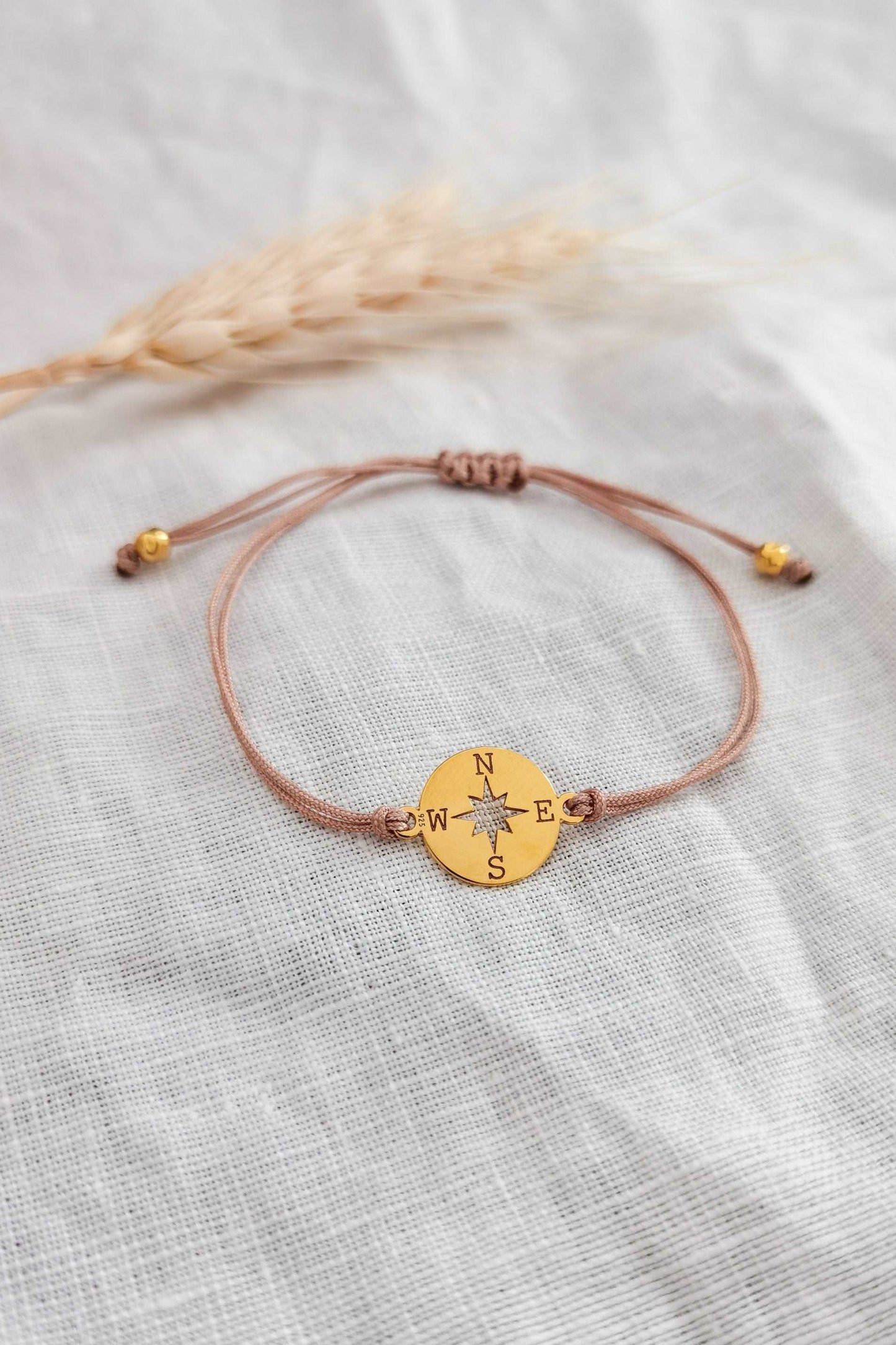 Goldenes Kompass Armband mit beigem Band vor weißem Hintergrund